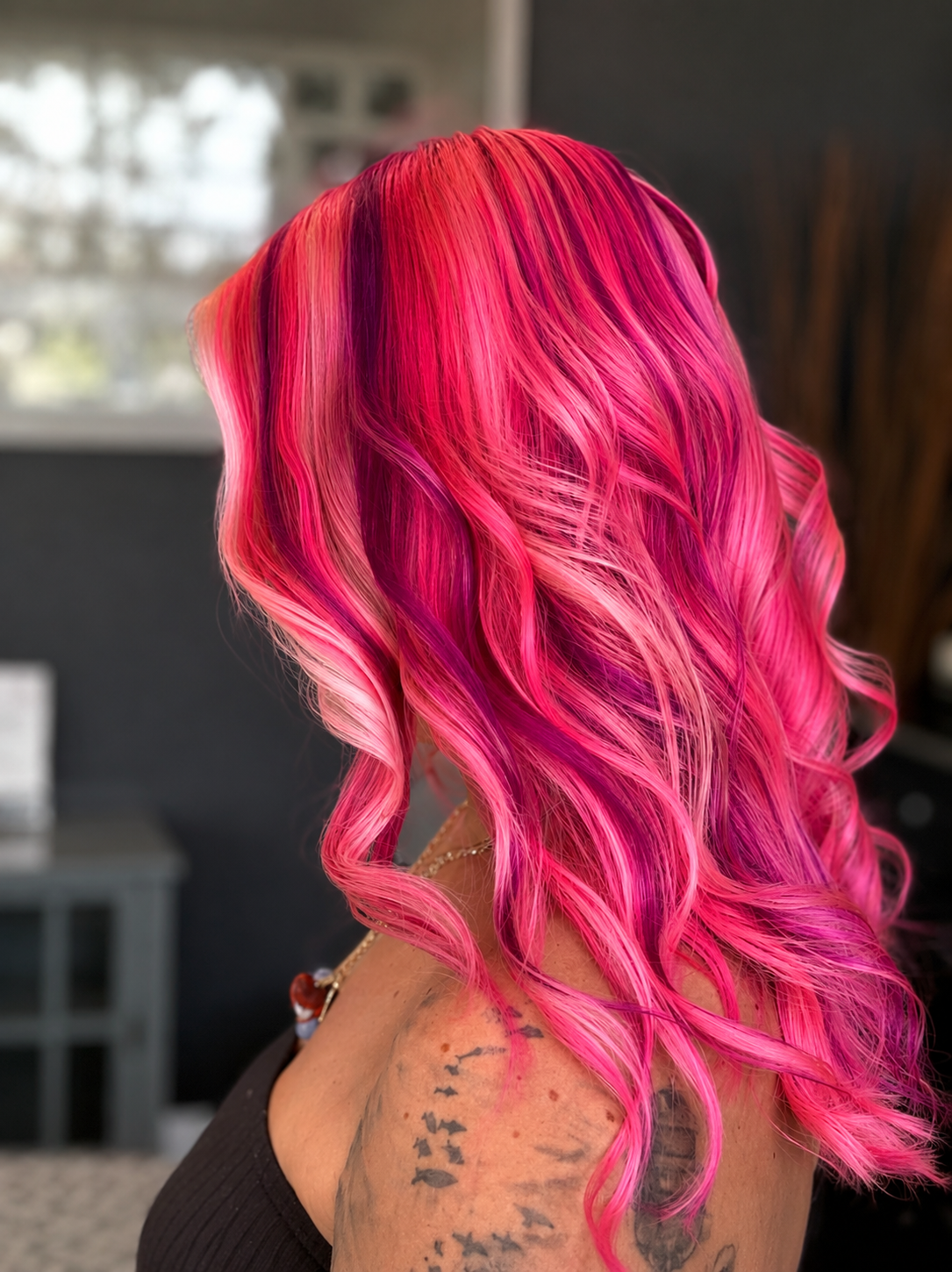 Side view of a woman with long, curly hair dyed in shades of pink and red, with tattoos on her shoulder and arm, in an indoor setting.