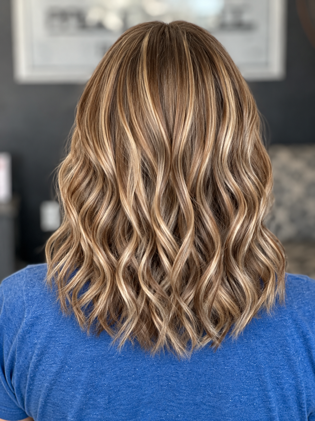 Back of a person's head showing shoulder-length, wavy blonde hair with highlights, wearing a blue shirt.