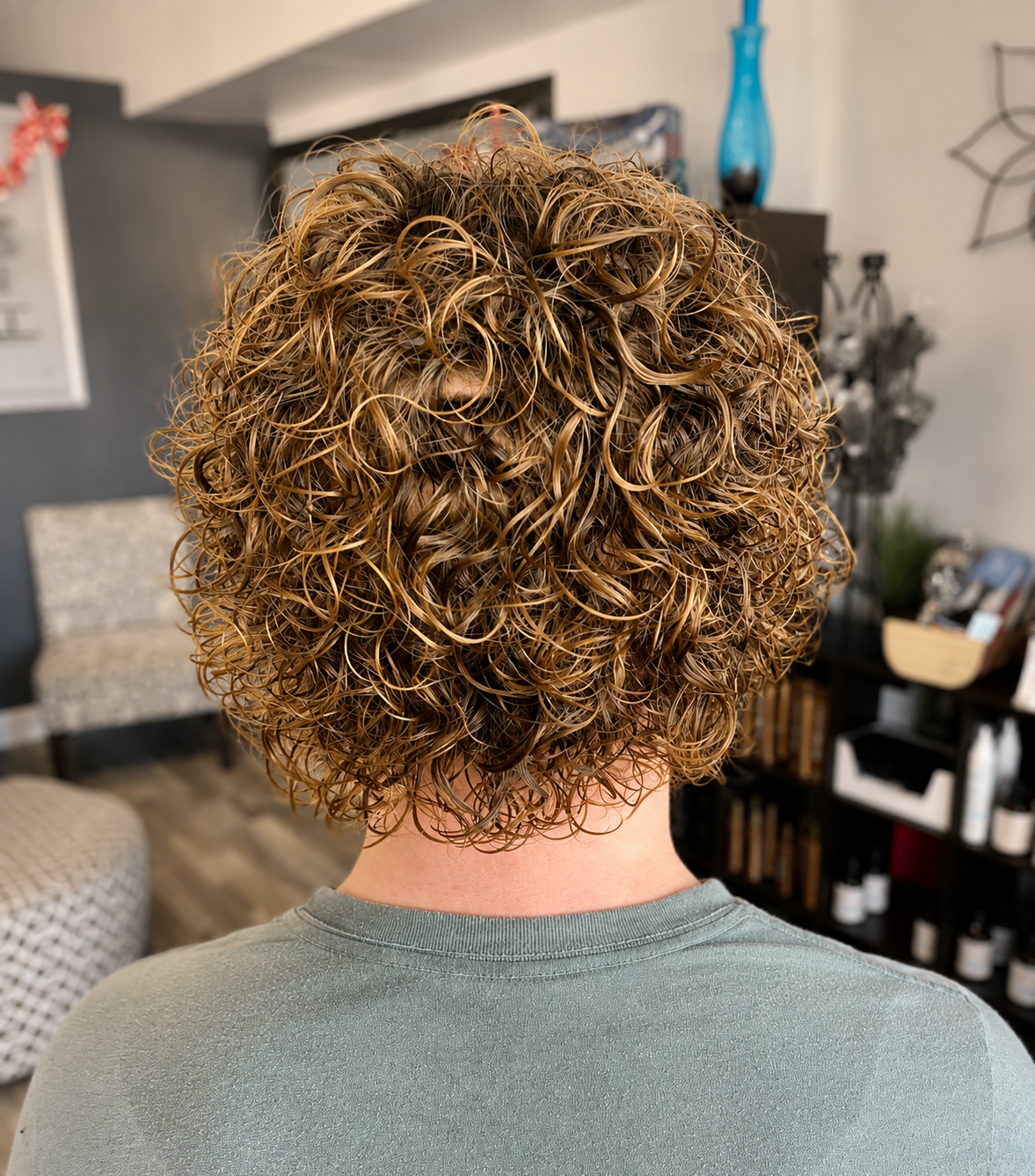 Back view of a person with short, curly, brown hair, wearing a gray shirt, in a room with various decorations and furniture.