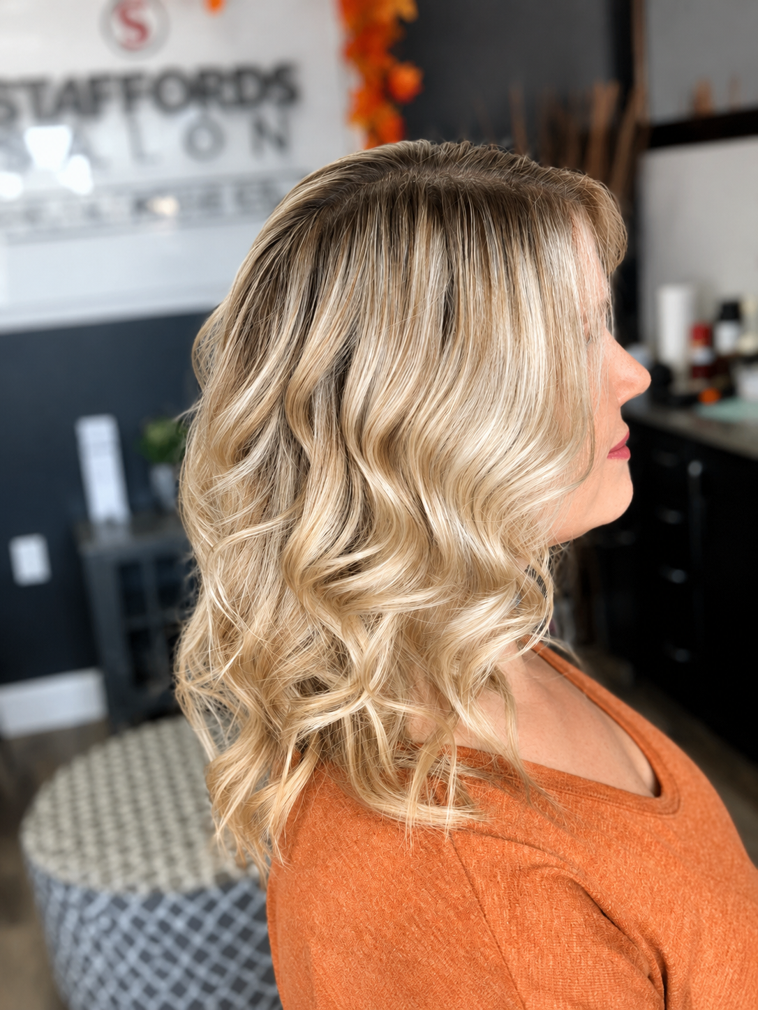 A woman with blonde, wavy hair wearing an orange top, seen in profile at a hair salon.