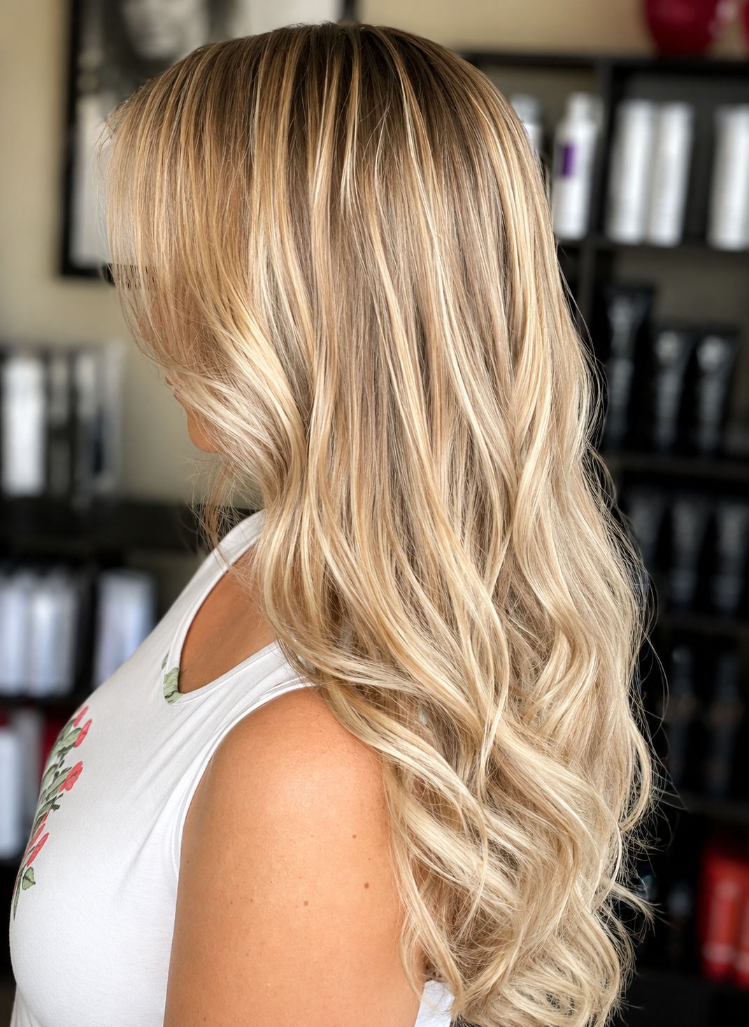 Side view of a woman with long, wavy blonde hair in a salon or store, with shelves of hair products in the background.