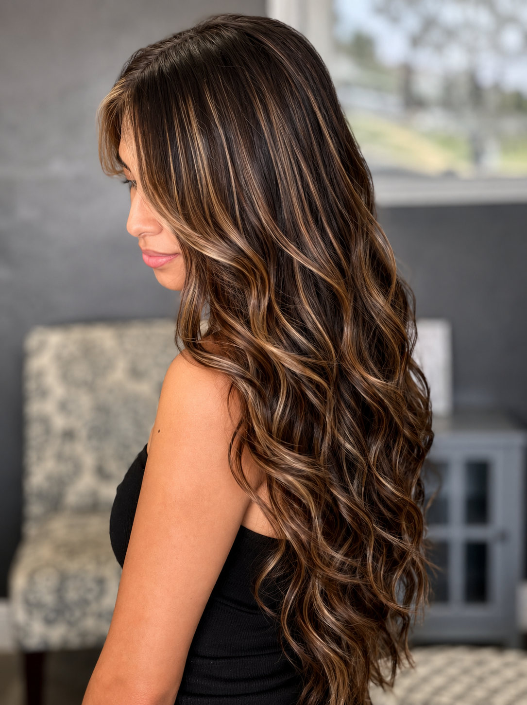 A woman with long, wavy brown hair with highlights, looking down in a black sleeveless top, in a modern indoor setting.