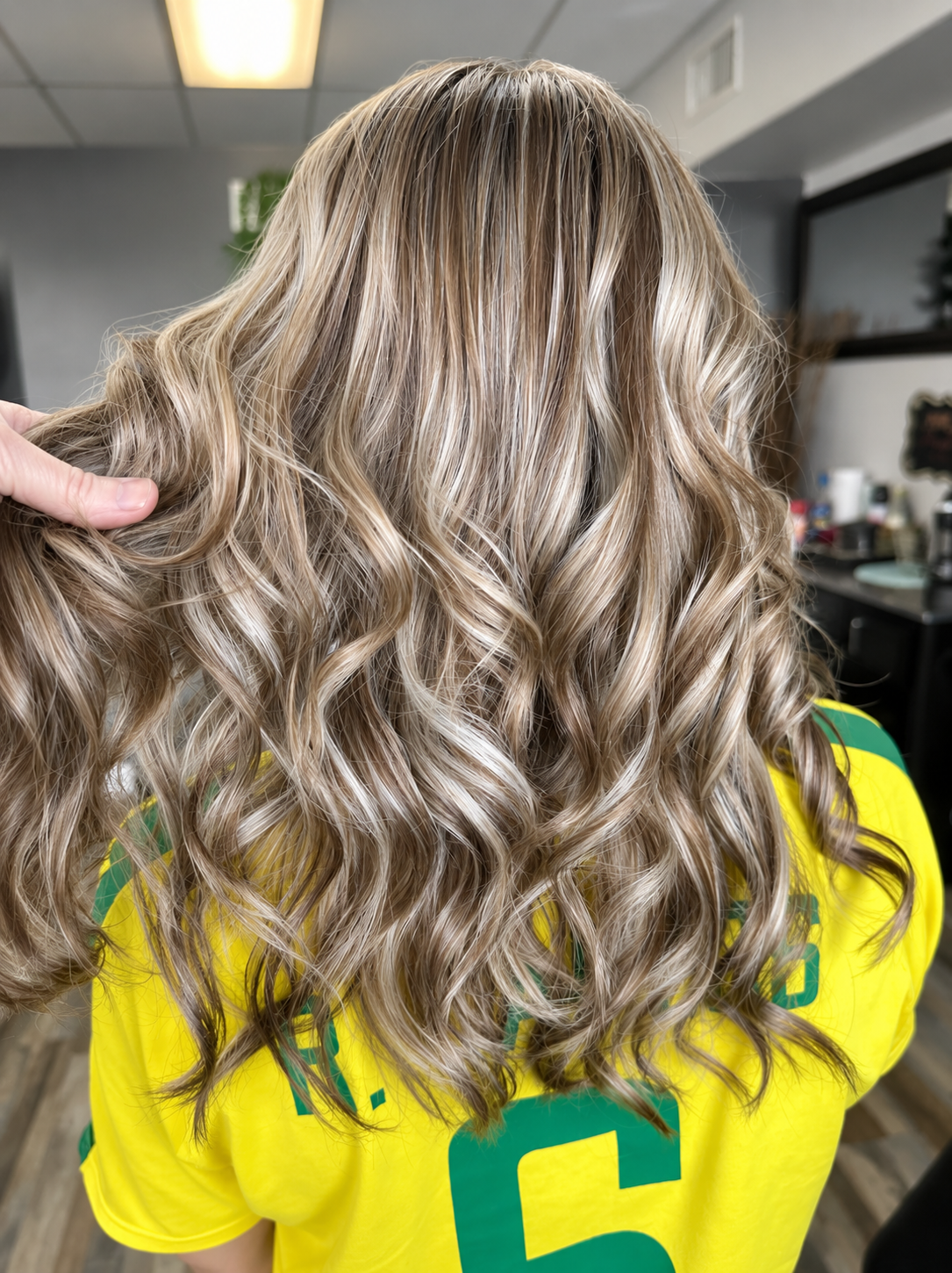 Close-up of a woman with long blonde, curly hair, wearing a yellow and green sports jersey, sitting in a salon or barbershop.
