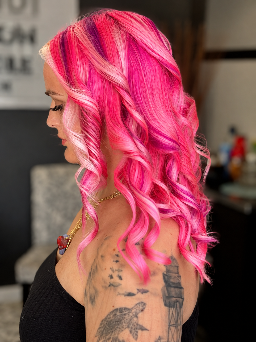 A woman with bright pink, curly hair and tattoos on her shoulder, wearing a black top and gold jewelry.
