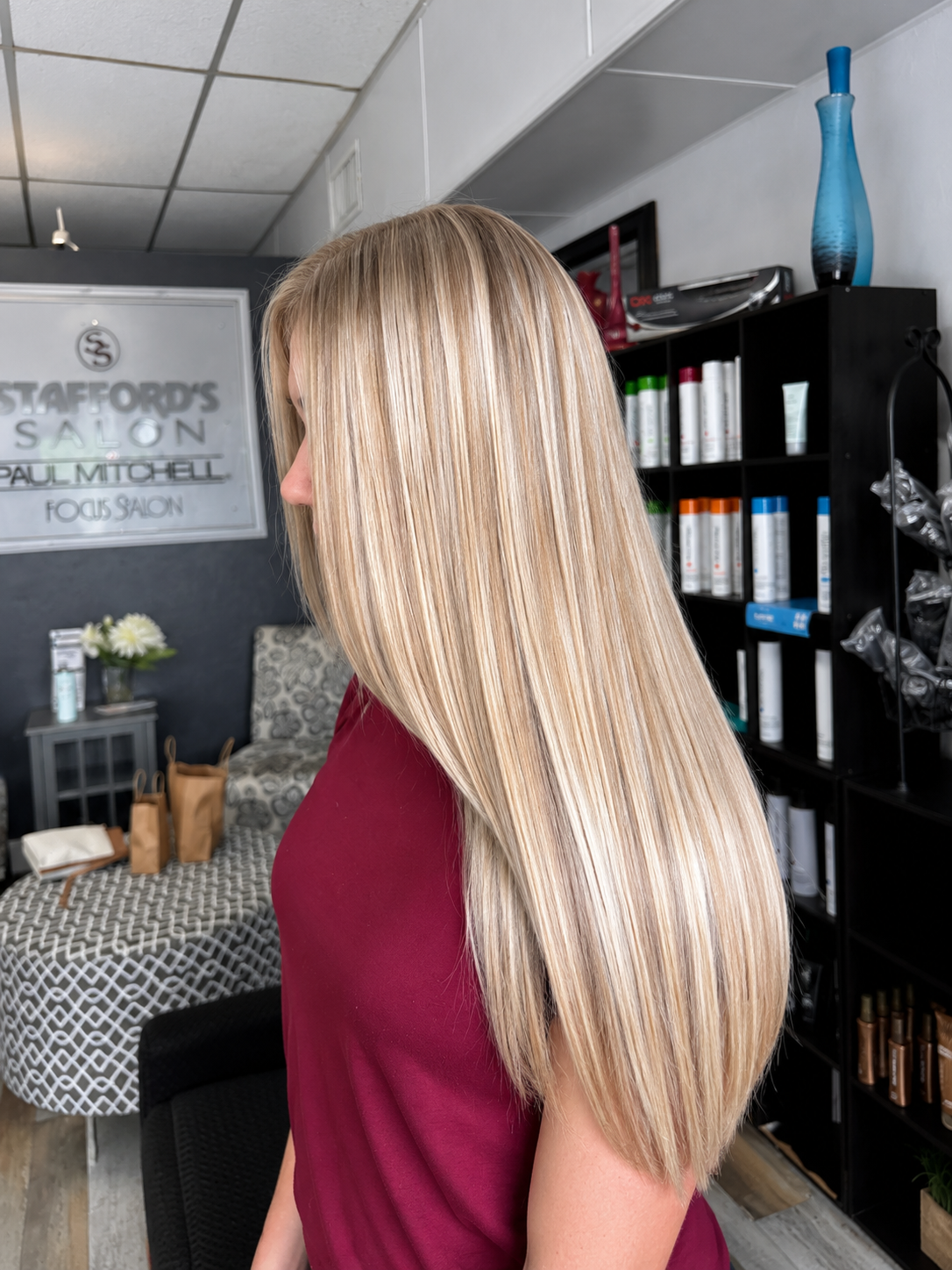 A woman with long, straight blonde hair wearing a maroon top sitting in a salon.