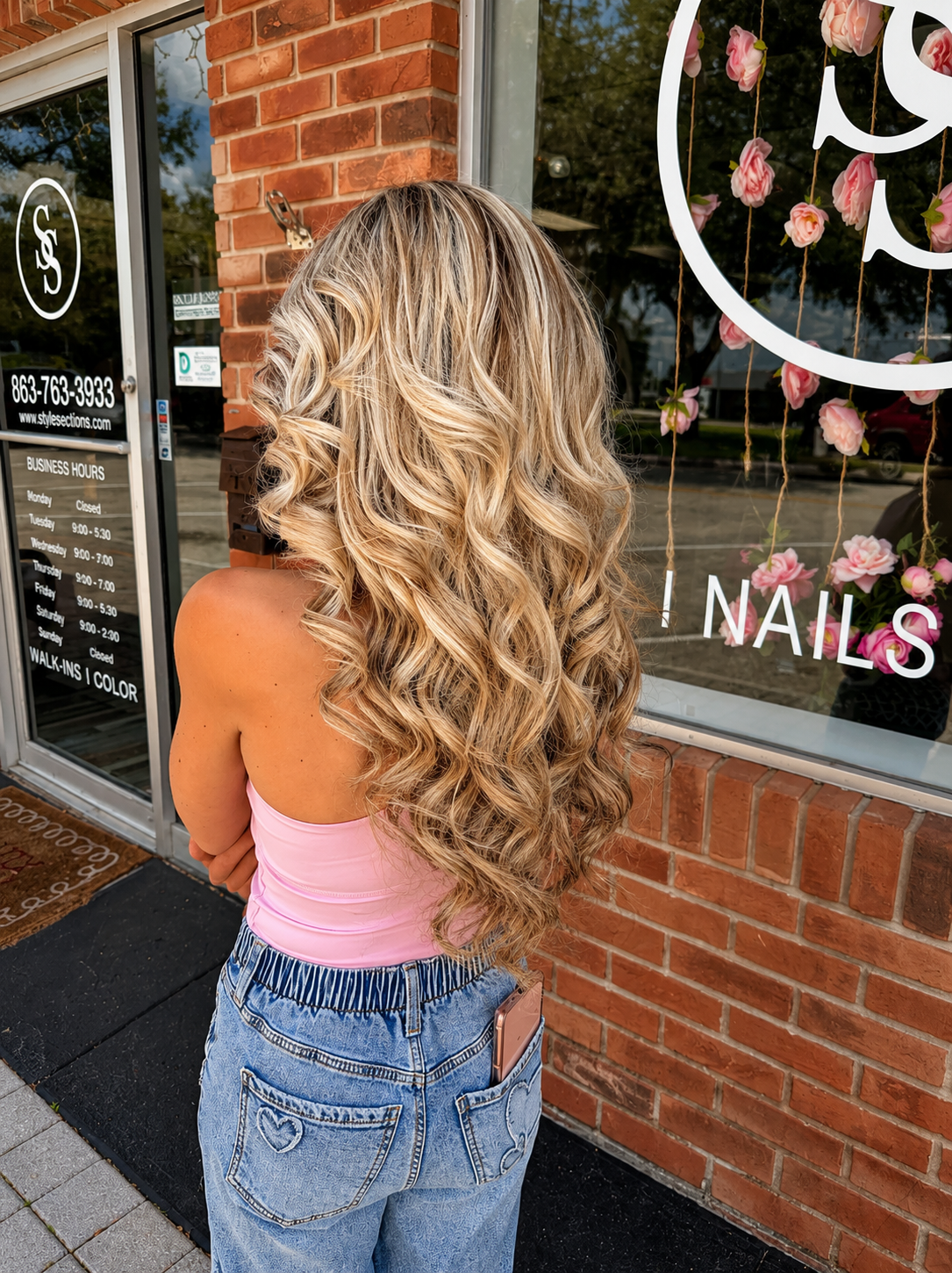 A woman with long, blonde, curly hair stands outside a nail salon, facing away. She wears a pink strapless top and high-waisted jeans with heart-shaped stitching on the back pockets. The salon window displays pink flowers and the text 'NAILS'.