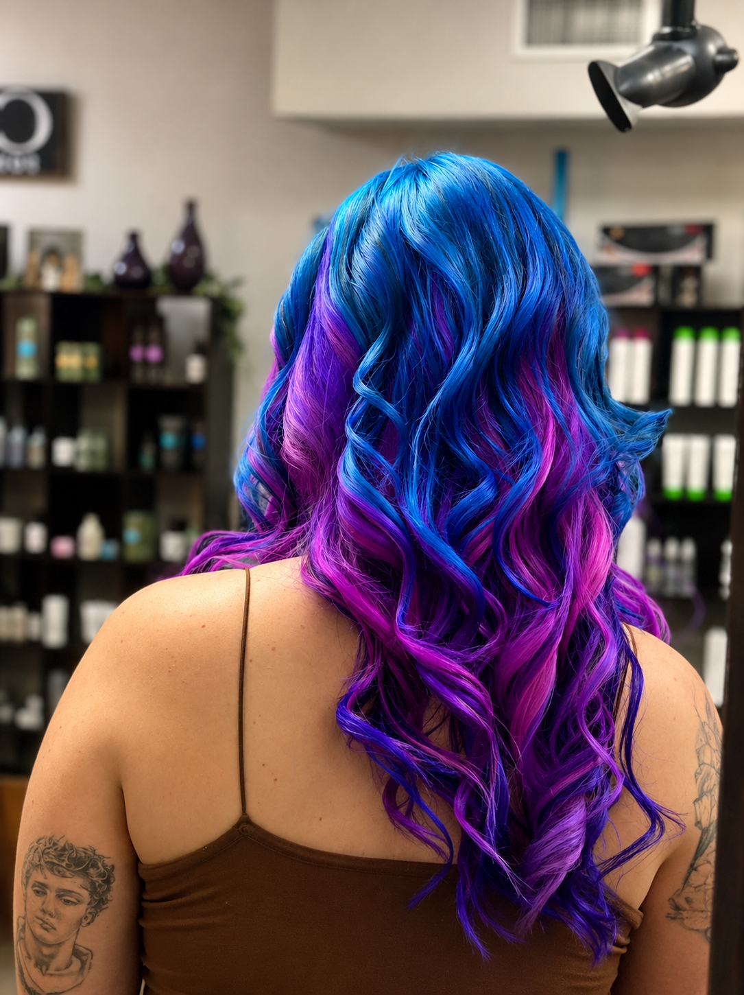 Back view of a woman with vibrant, curly hair dyed in shades of blue, purple, and pink, in a salon setting.