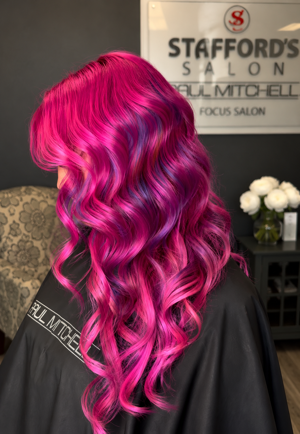 Woman with long, vibrant pink and purple wavy hair at a salon, with a sign for Stafford's Salon in the background.