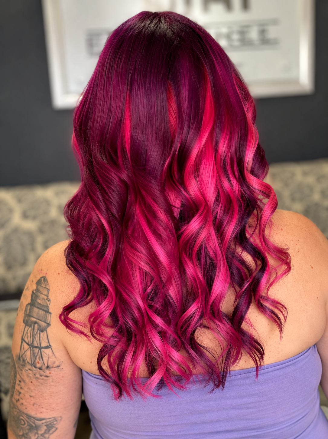 Woman with long, curly hair dyed in shades of pink and purple, wearing a purple strapless top, with tattoos on her arm, in a salon or studio setting.