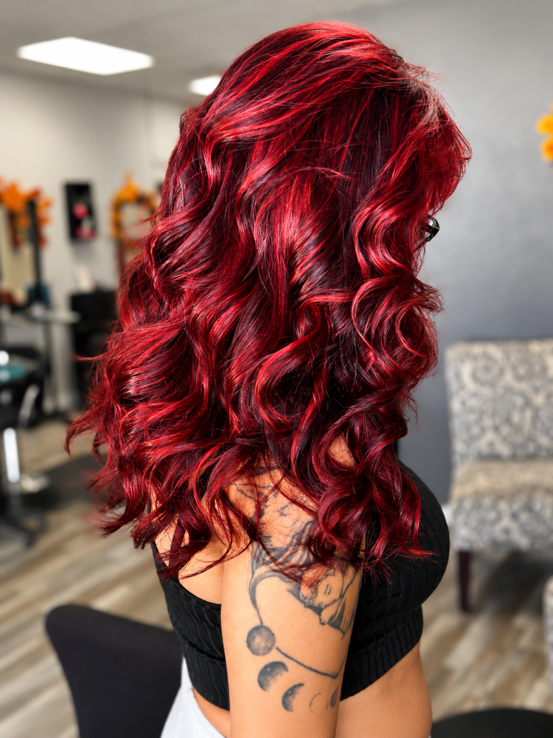 The image shows a woman with long, wavy, red hair sitting in a salon. She has a tattoo on her upper arm and is wearing a black top. The background includes salon mirrors and chairs.