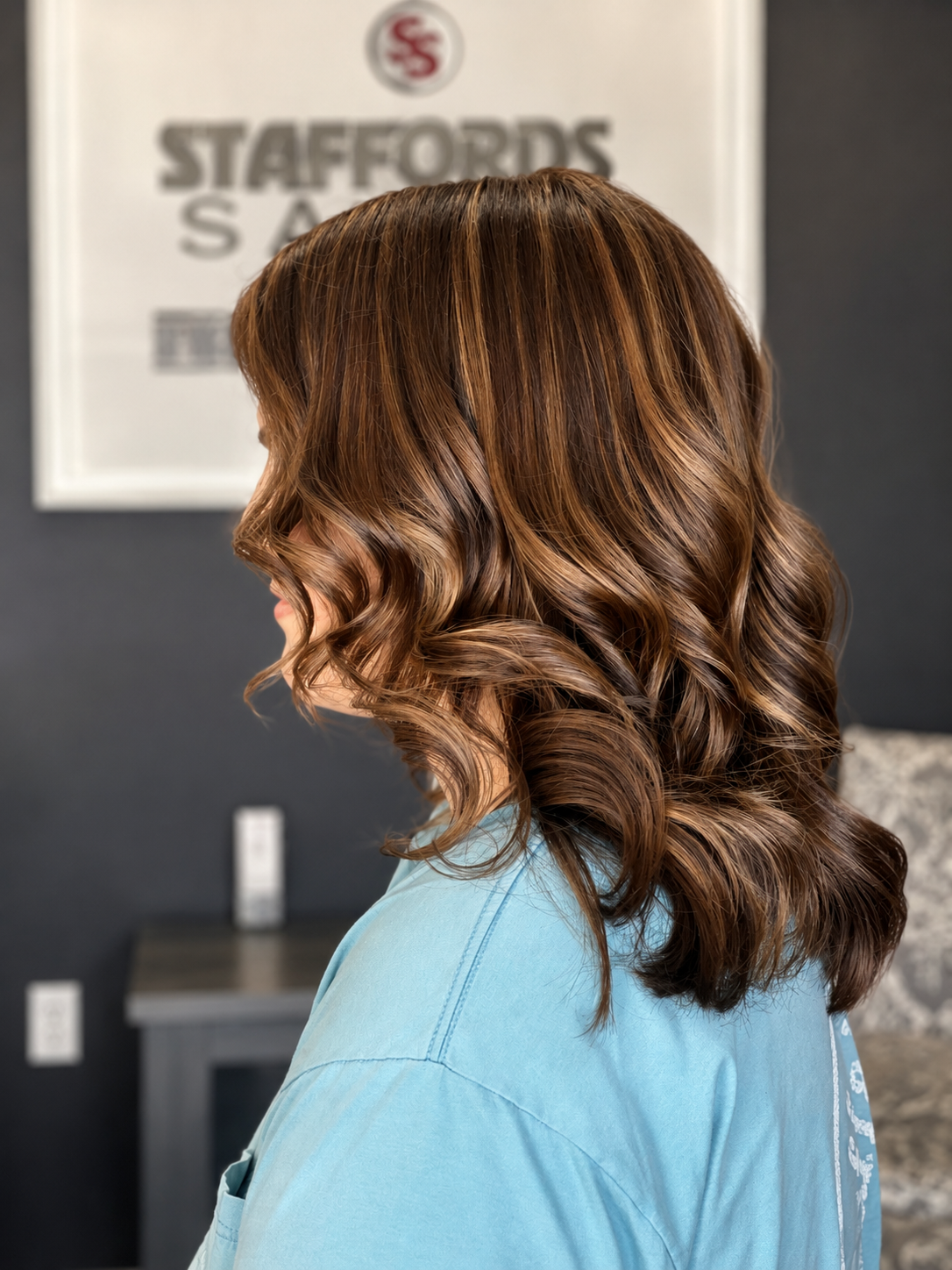 Profile view of a woman with medium-length, wavy, caramel-colored hair, wearing a light blue shirt, in a salon with a 'Staffords Amb' poster in the background.