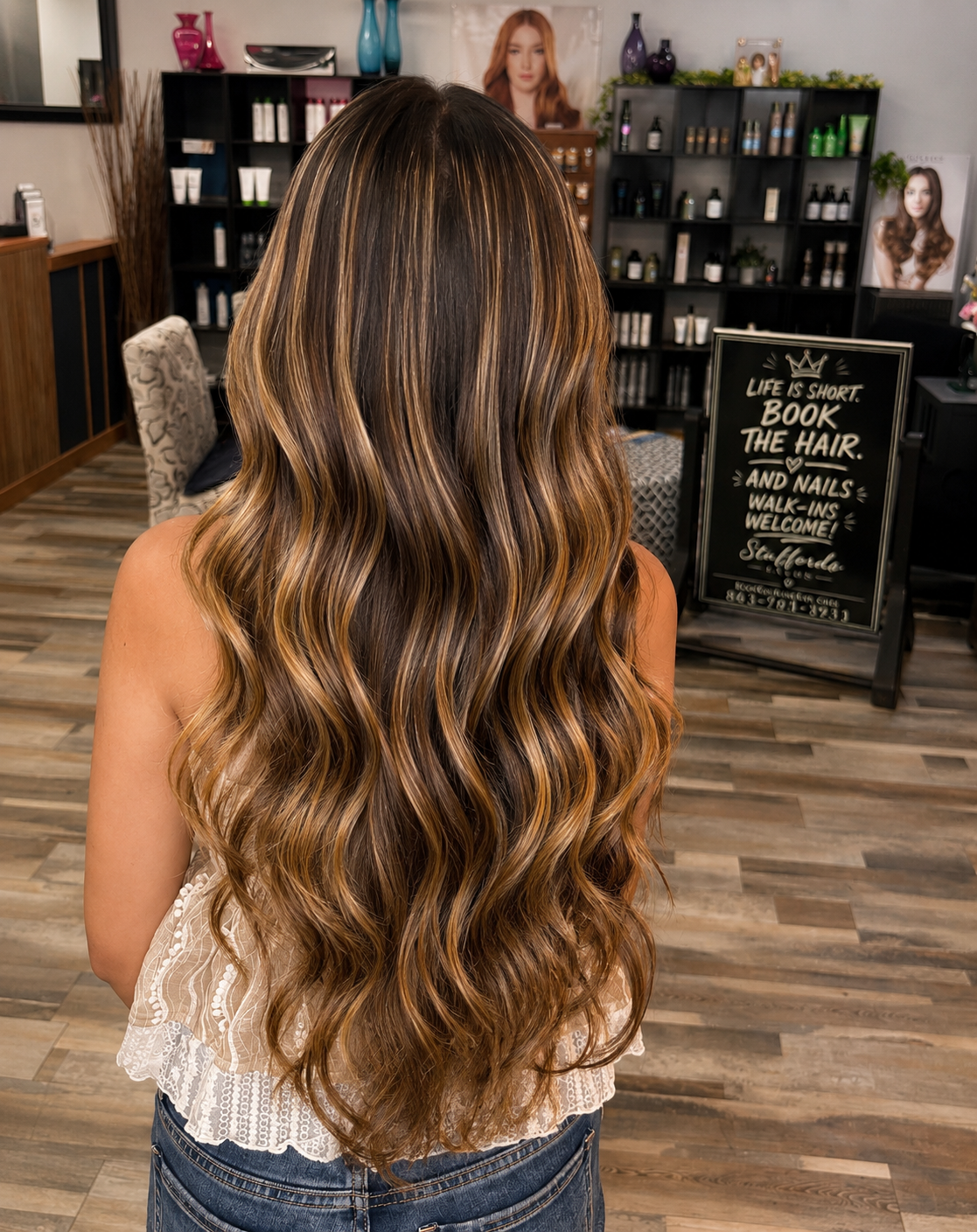 Back view of a woman with long, wavy, caramel-colored hair with blonde highlights inside a hair salon.