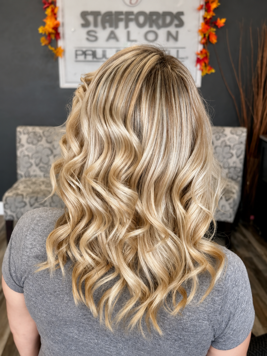 Back view of a woman with long, blonde, wavy hair sitting in a salon with a sign reading 'Staffords Salon' in the background.