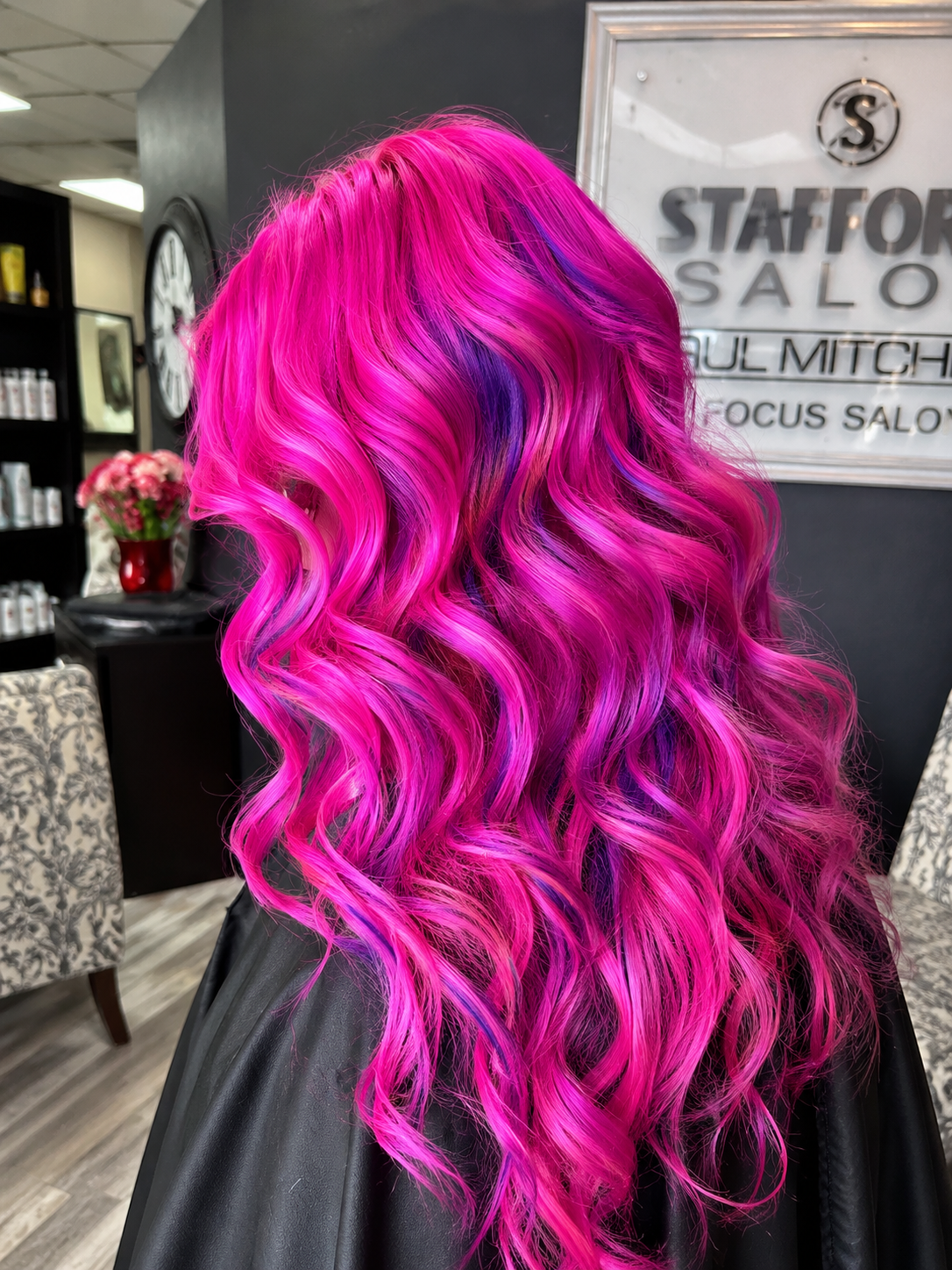 Back view of a person with long, vibrant pink and purple curly hair inside a salon.