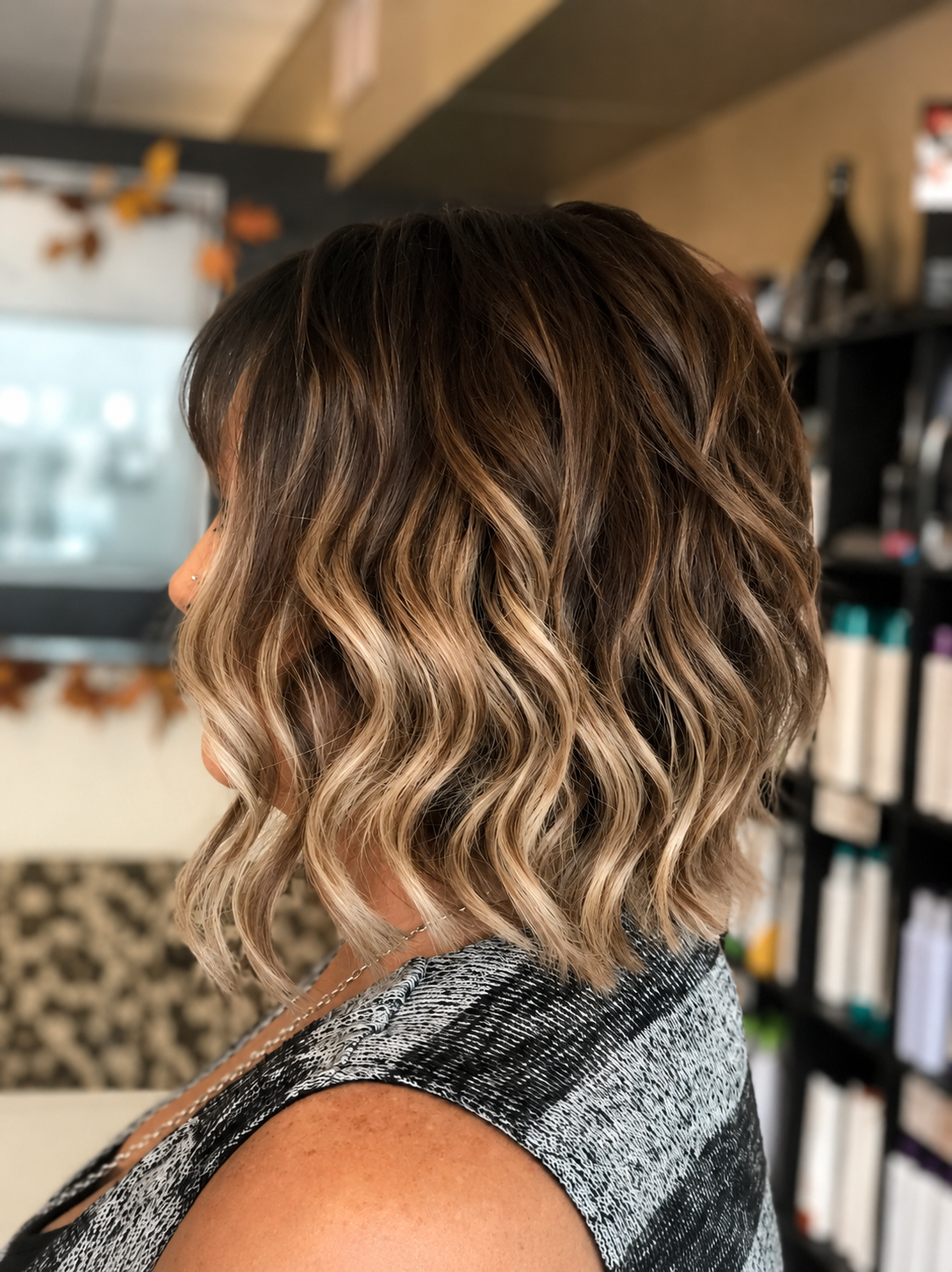 A woman with shoulder-length wavy hair that transitions from dark brown at the roots to blonde highlights at the ends, standing in a hair salon.