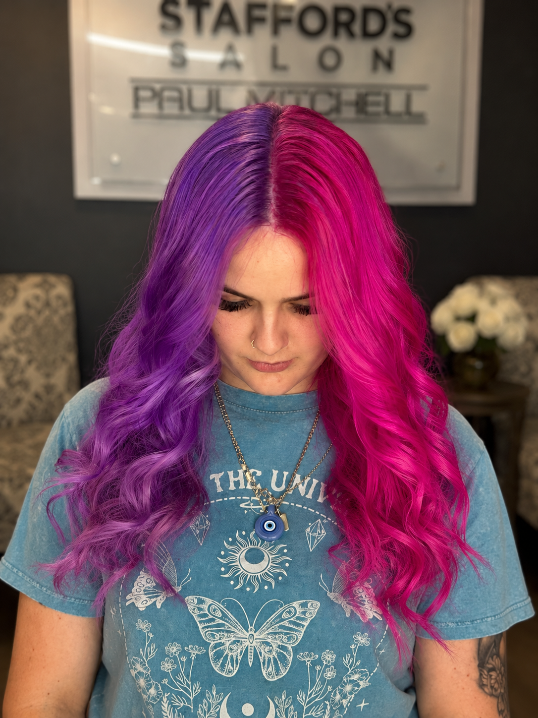 A woman with long, wavy hair dyed in vibrant purple and pink shades, looking down. She is wearing a blue graphic t-shirt with butterfly and celestial designs, layered necklaces including an evil eye pendant, and has a nose ring. She is in a salon wit