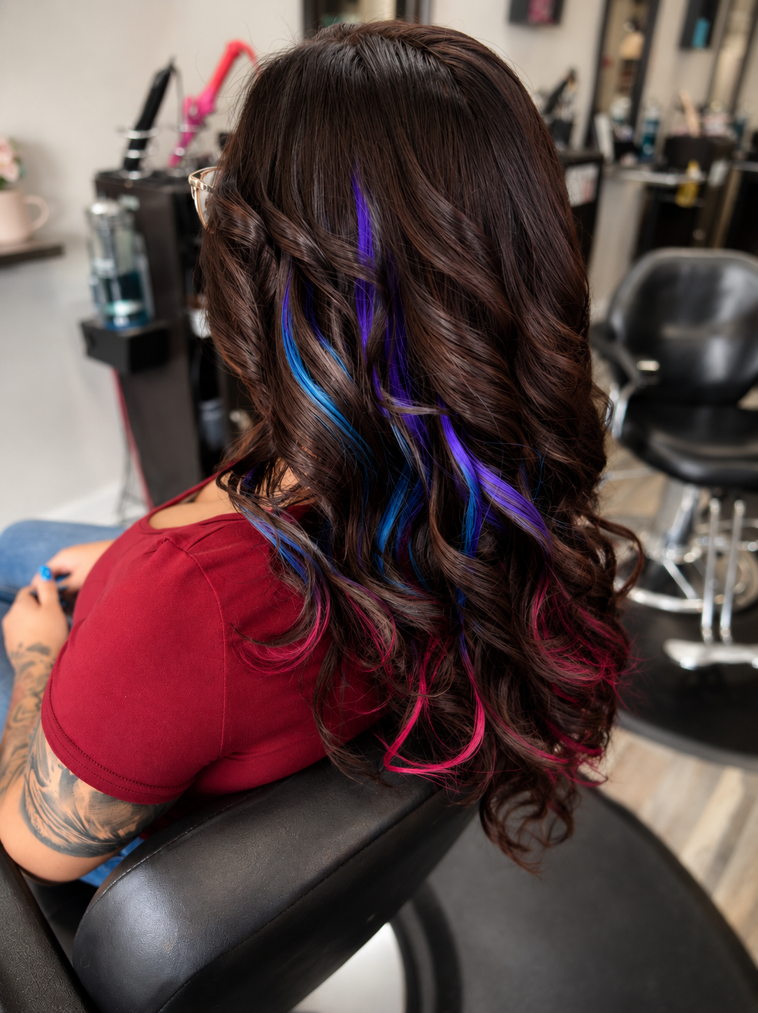 A woman with long, dark brown hair styled in loose curls, with vibrant blue and purple streaks, sitting in a salon chair with her back to the camera.