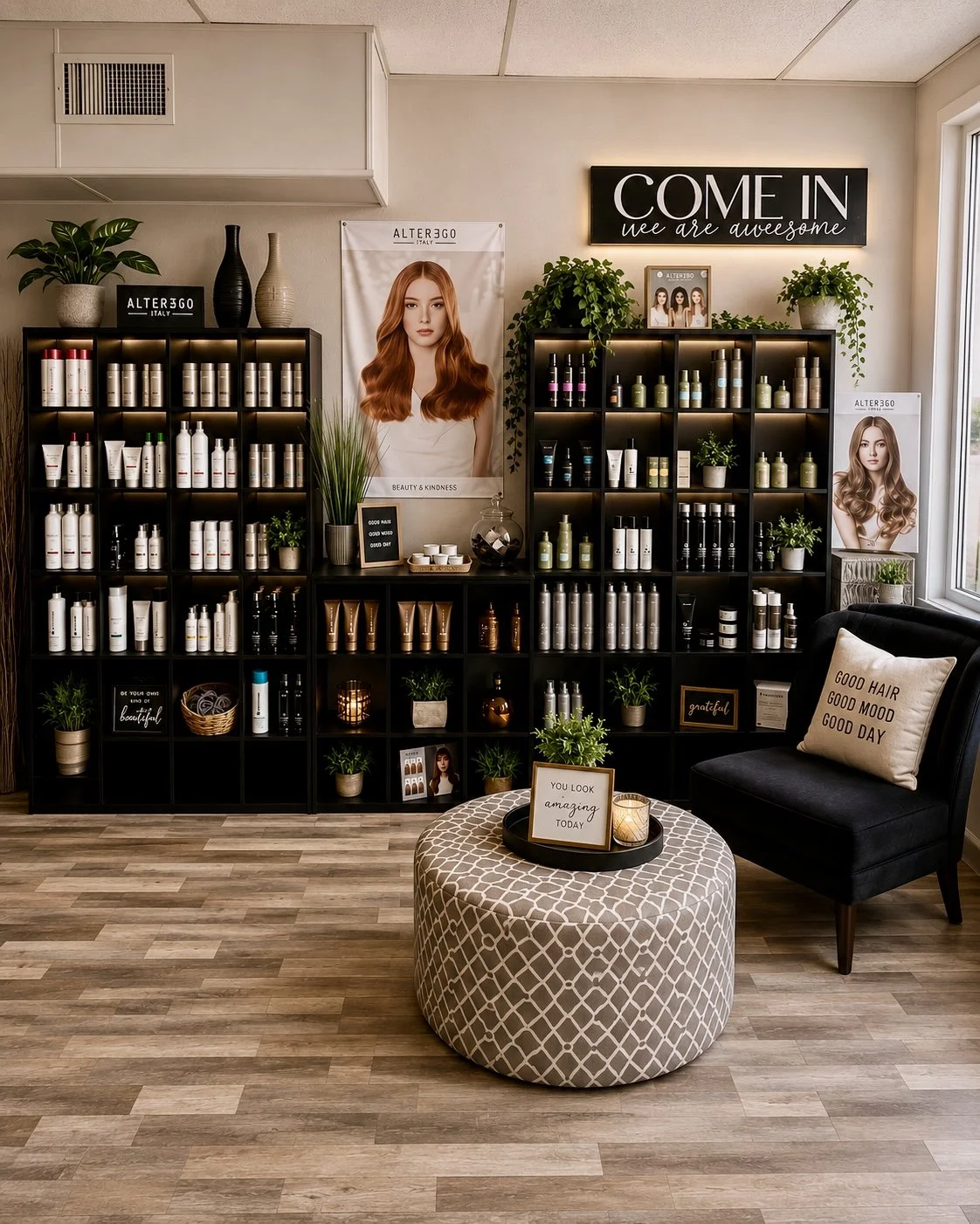Interior of a beauty or hair salon featuring black shelving units with hair products, potted plants, posters of women with styled hair, a cozy seating area with a pillow reading 'Good Hair Good Mood Good Day,' and decorative accents like signs and ca