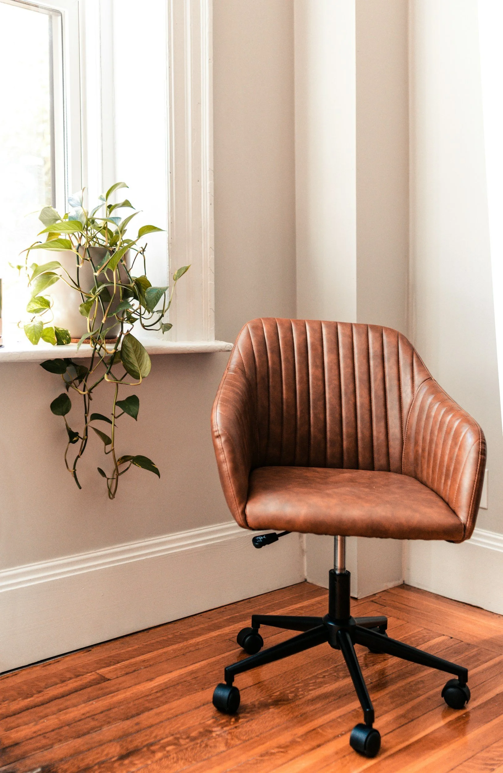 A brown leather office chair on casters placed on a hardwood floor, beside a white wall, with a window sill holding a green potted plant, and beige walls in the background.