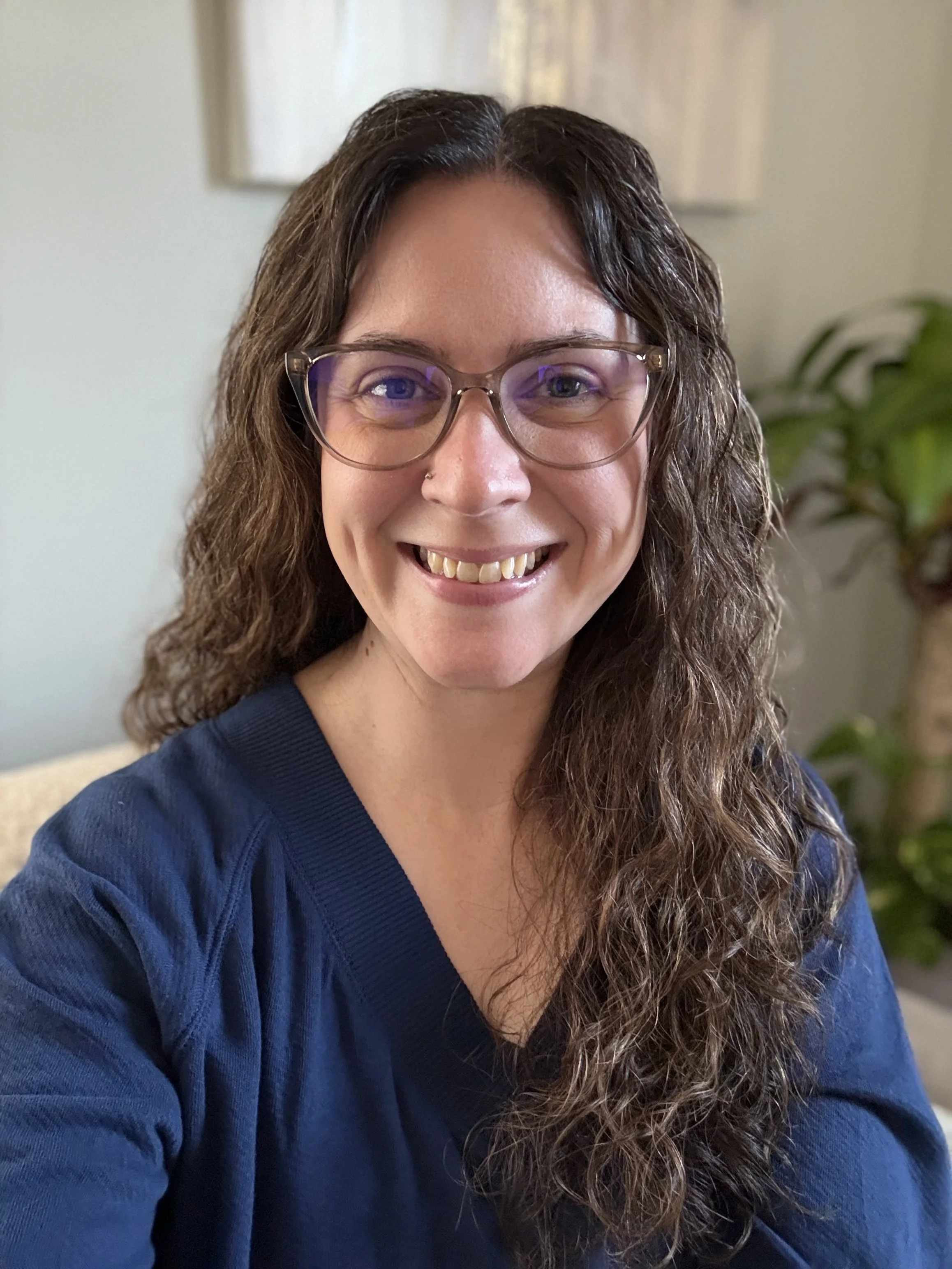 A woman with curly brown hair, glasses, and a nose ring smiling while wearing a blue sweatshirt, indoors with a plant and wall in the background.