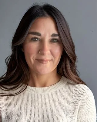 Close-up portrait of a woman with dark brown hair, wearing a nose ring and a light-colored sweater, smiling slightly against a plain gray background.