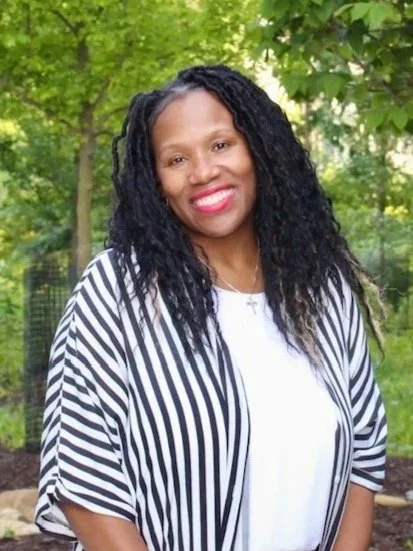 Kim Lott hair smiling outdoors with green trees in the background, wearing a white top and a black and white striped jacket.