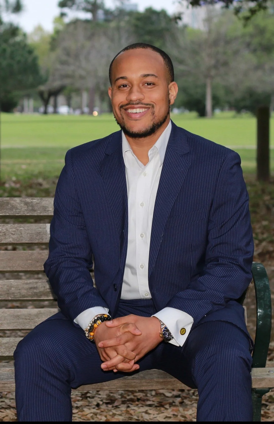 Ja'Von Long wearing a navy pinstripe suit and white dress shirt sitting on a park bench, smiling with hands clasped, in an outdoor park with green grass and trees in the background.