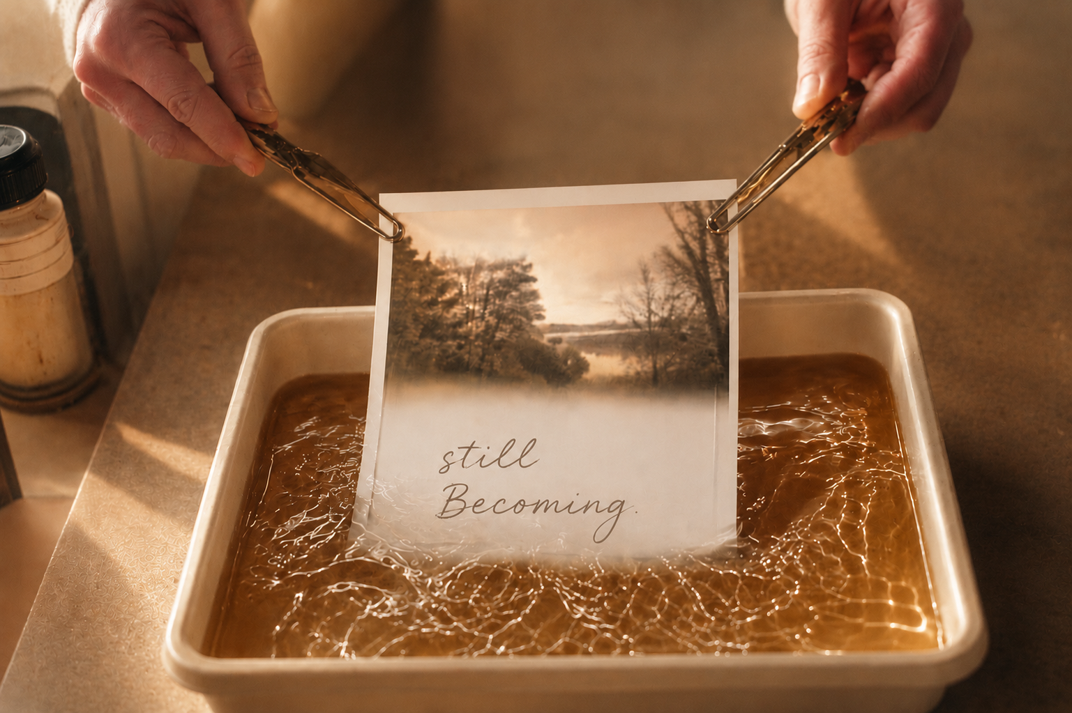A person is placing a photograph with the words 'still Becoming' in a tray of water, creating a reflection. The photograph depicts a landscape with trees and a body of water.