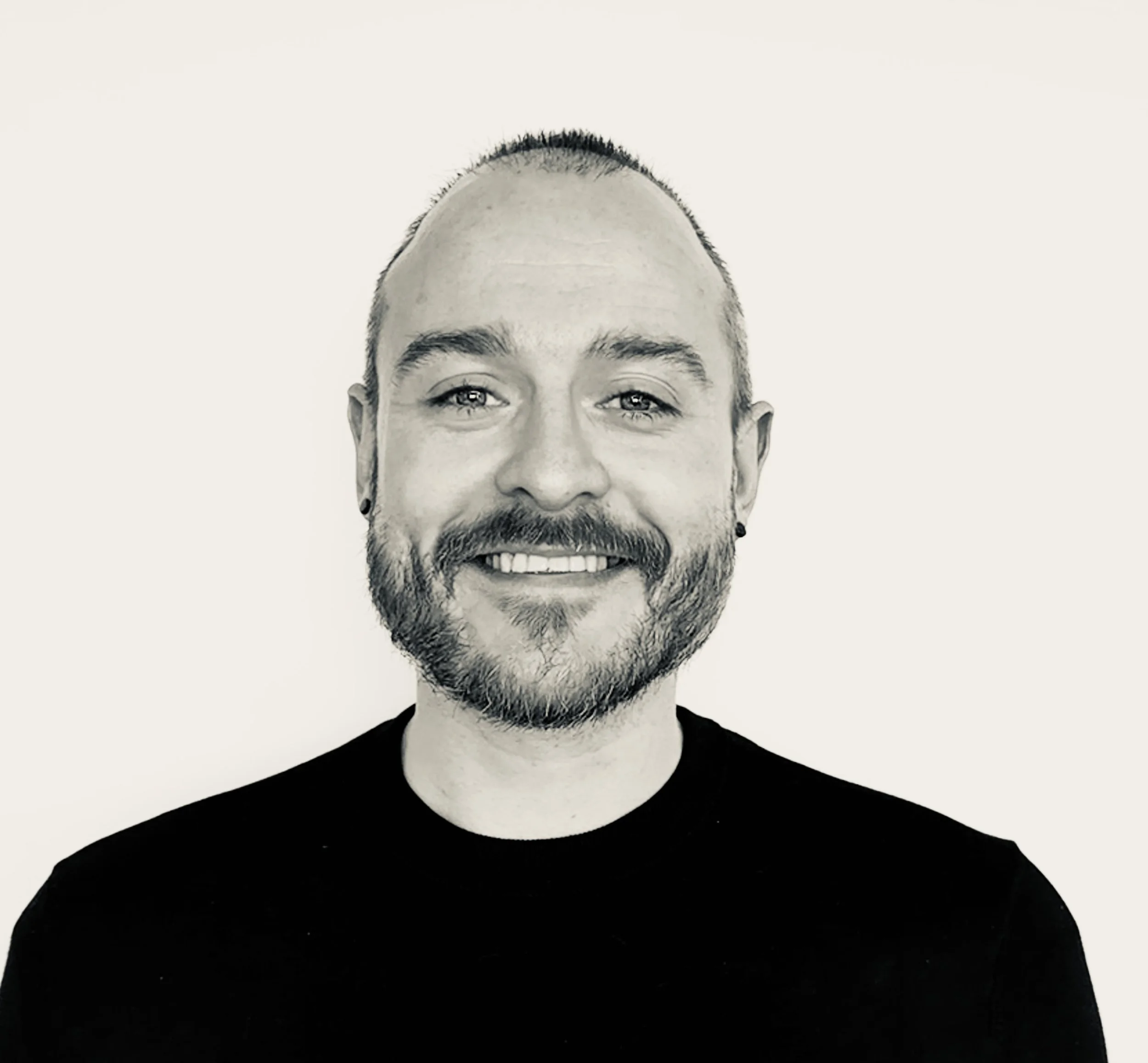 A black and white digital photo of a smiling man with short hair and a beard, wearing a black shirt, against a plain light background.