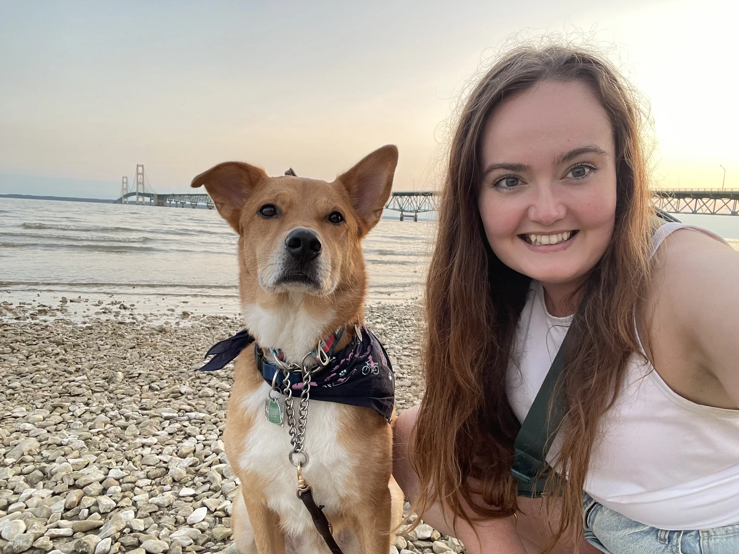 A young woman with long brown hair smiling alongside a tan and white dog at the beach near a large bridge in the background during sunset.