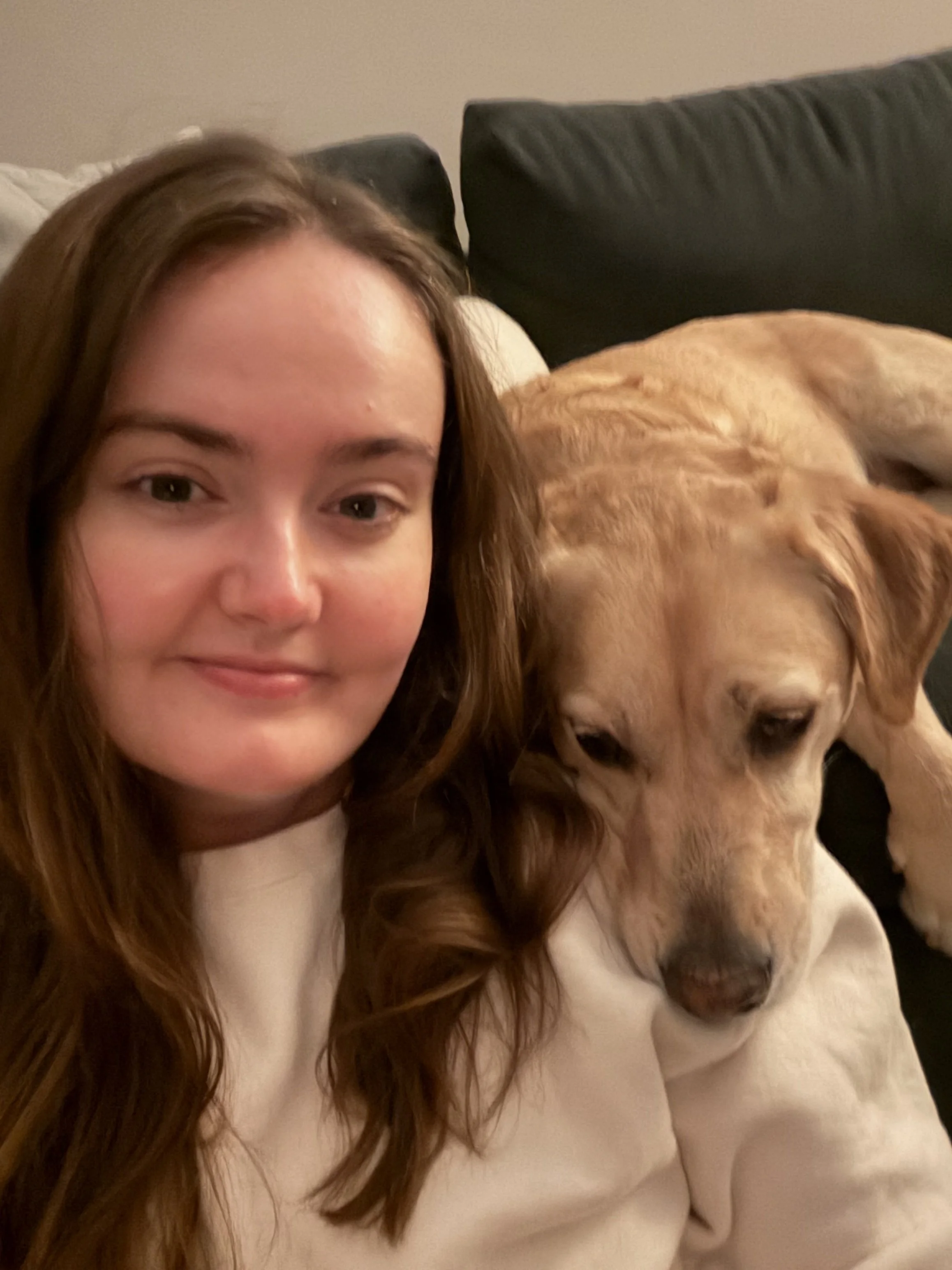 A woman with long brown hair and fair skin sitting on a beige couch, next to a large golden retriever dog with light golden fur.