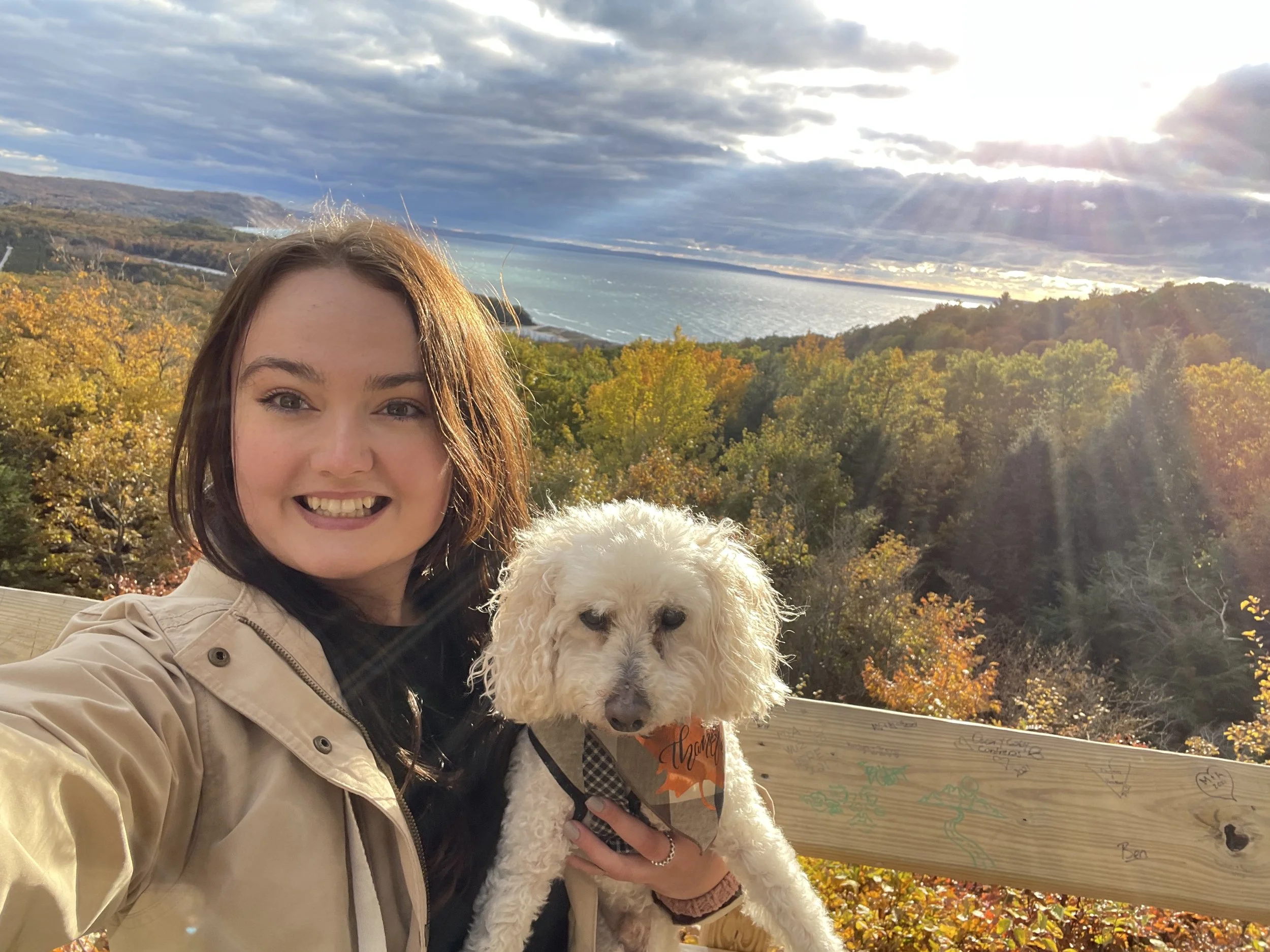A smiling woman holding a white fluffy dog with a checkered scarf, in front of an artistic wooden panel with colorful drawings, overlooking a scenic landscape with blue water, trees with autumn foliage, and a cloudy sky.