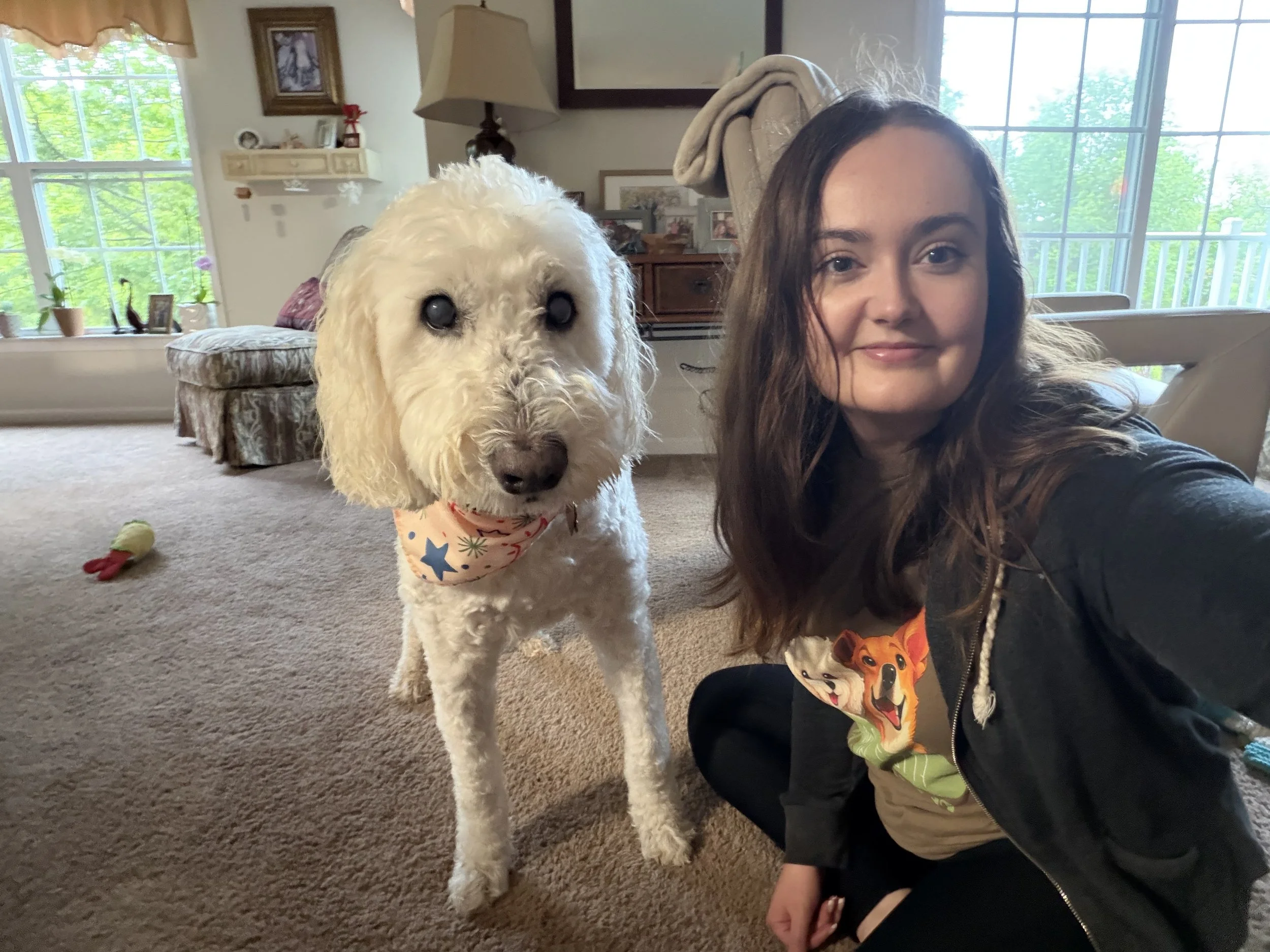A young woman with long brown hair taking a selfie with a large, curly-haired white dog. The dog is wearing a bandana with colorful stars, and the woman is smiling softly. They are in a living room with large windows showing green trees outside, a beige couch, and various decorations.