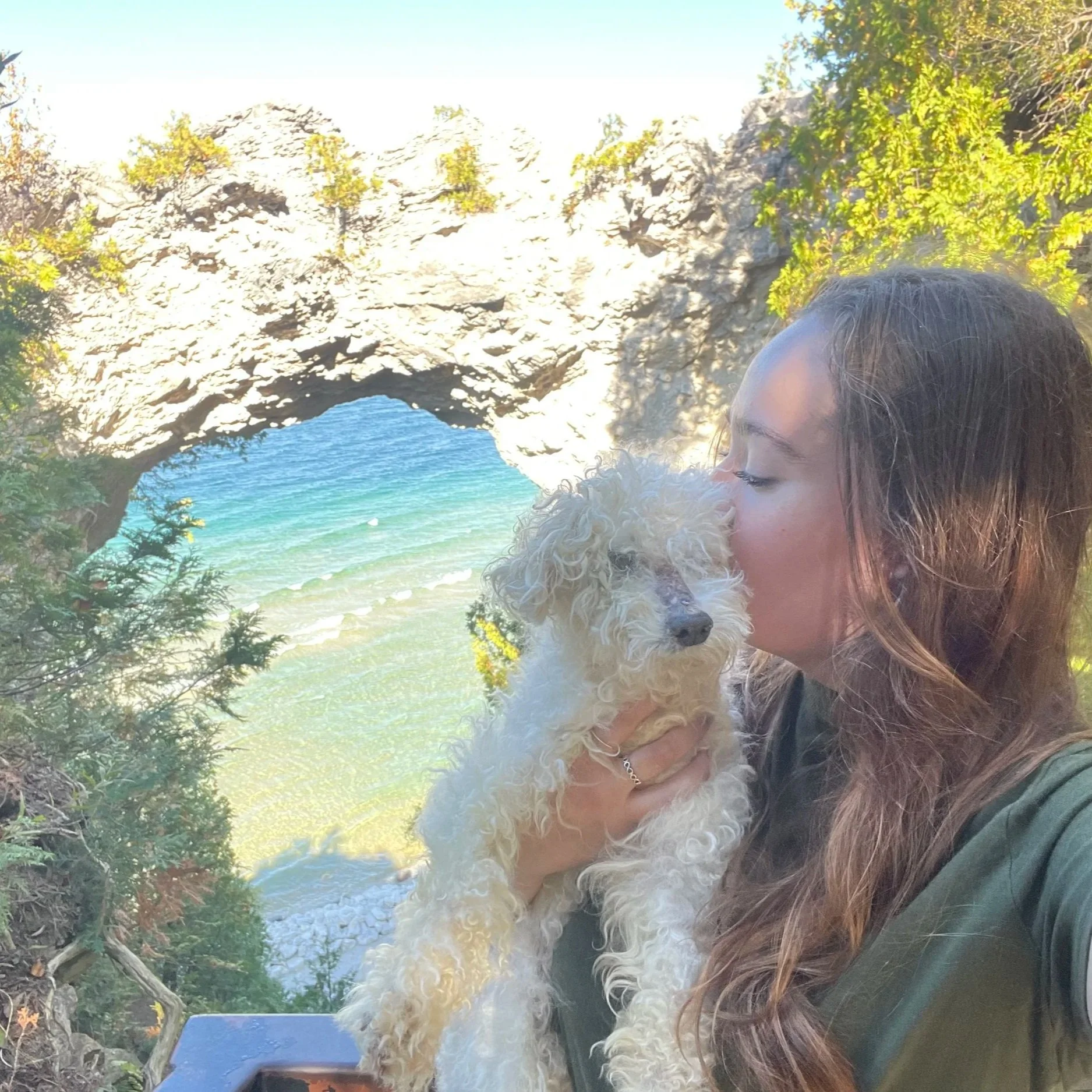 A woman with brown hair holding a curly white dog on a beach with rocky arch, trees, and ocean in the background.