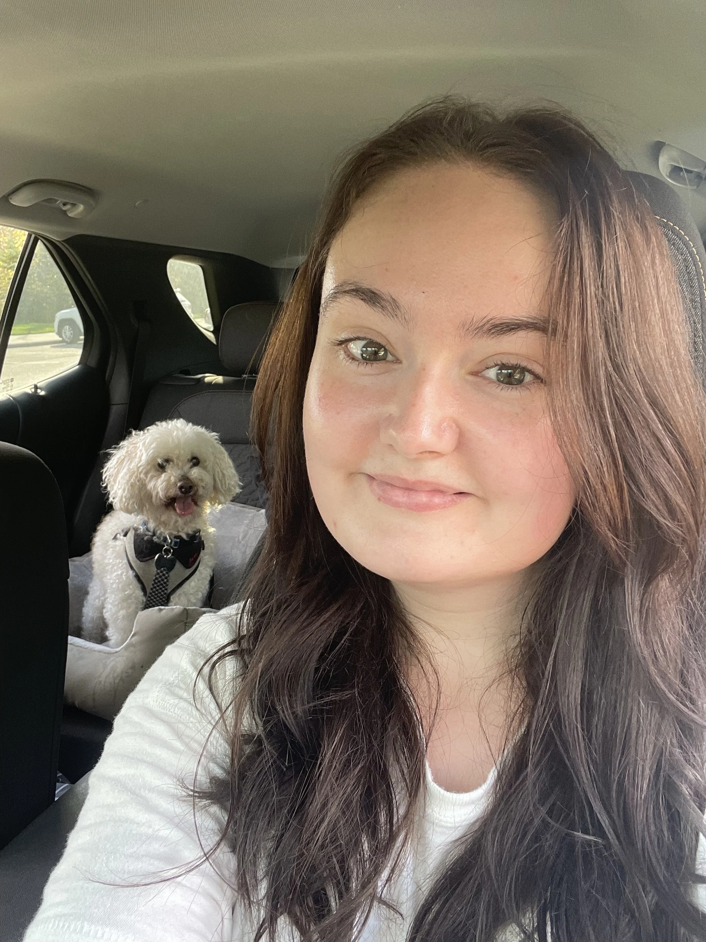 A young woman with long brown hair and hazel eyes taking a selfie inside a vehicle, with a small white curly-haired dog sitting on a tan pet booster seat in the background.