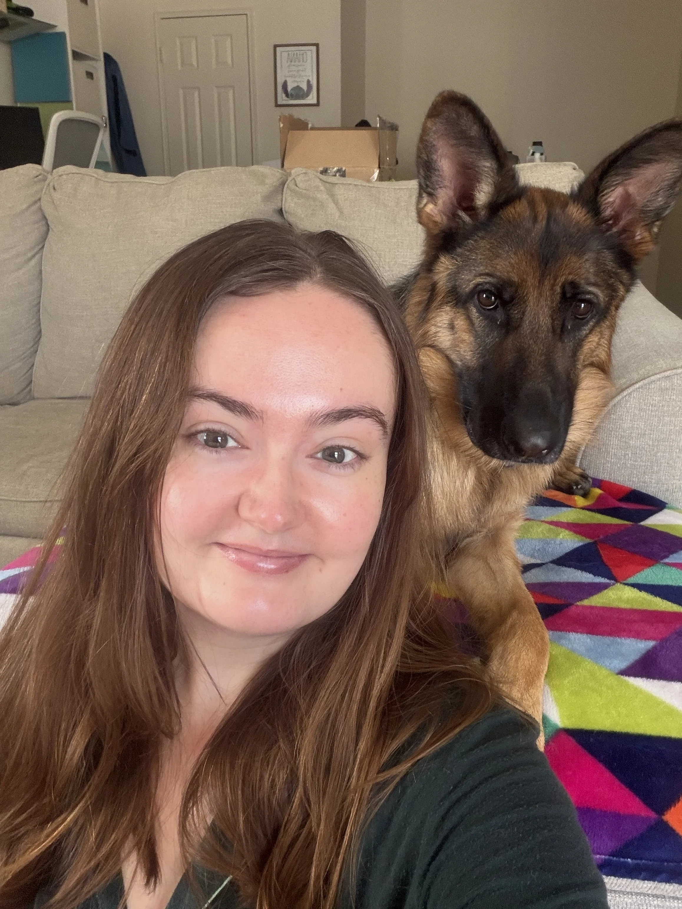 Close-up selfie of a woman with long brown hair and a friendly expression, sitting next to a large German Shepherd dog with alert ears, on a colorful blanket in a cozy living room.