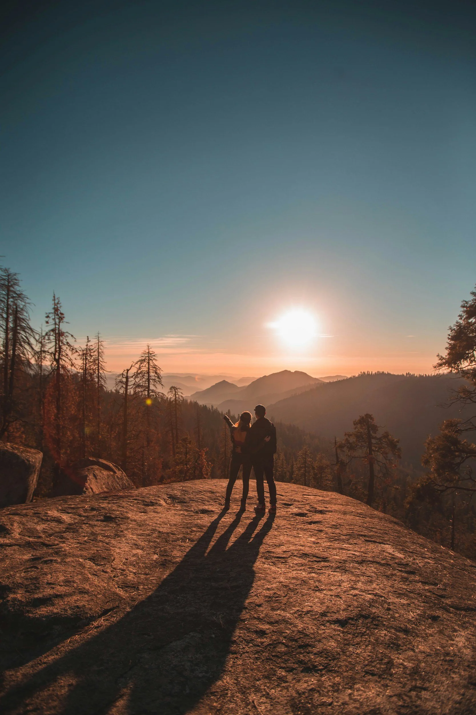 A couple standing on a rocky ledge at sunset, shadowed, overlooking a mountainous forest landscape with tall trees and distant mountain ridges.