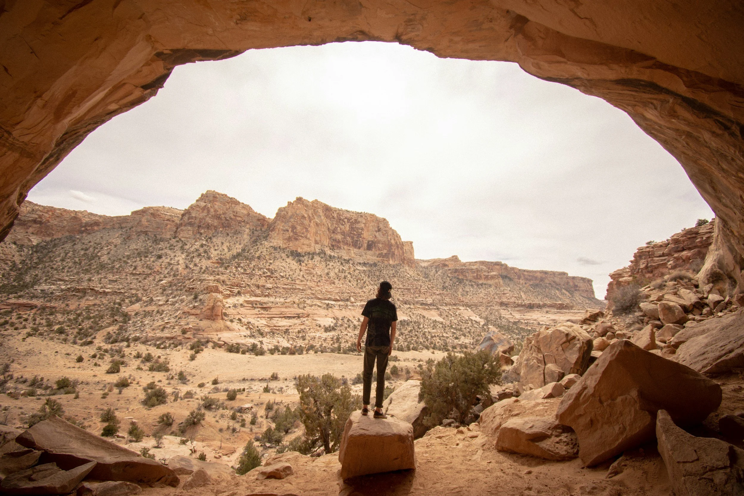 A person standing on a rock and looking out from a natural rock formation archway in a desert landscape with mountains and bushes.