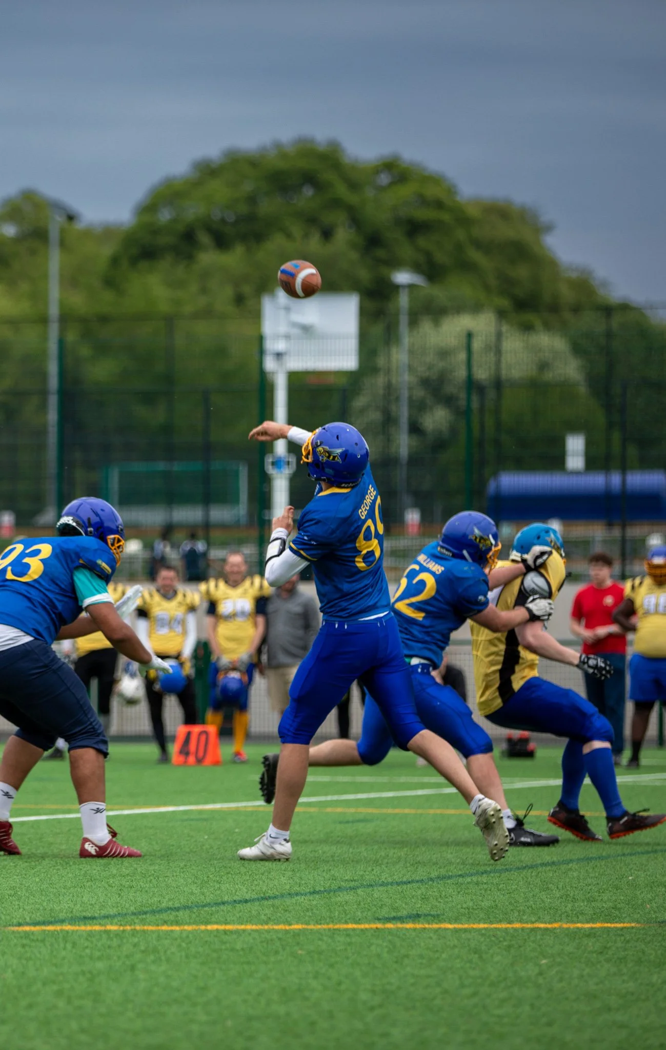 A football game in progress with players in blue and yellow uniforms, a player in blue jumping to catch the football, and officials and spectators watching. Overcast sky with green trees in background.
