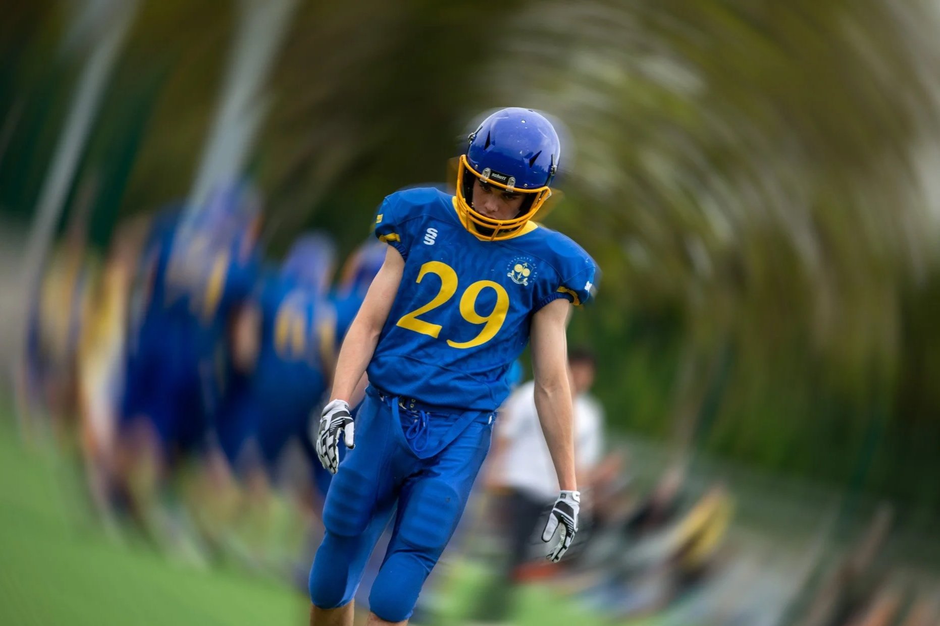 An American football player in a blue uniform with the number 29, wearing a blue helmet with a yellow faceguard, appears to be in a game or practice on a grassy field, with spectators or team members in the background.