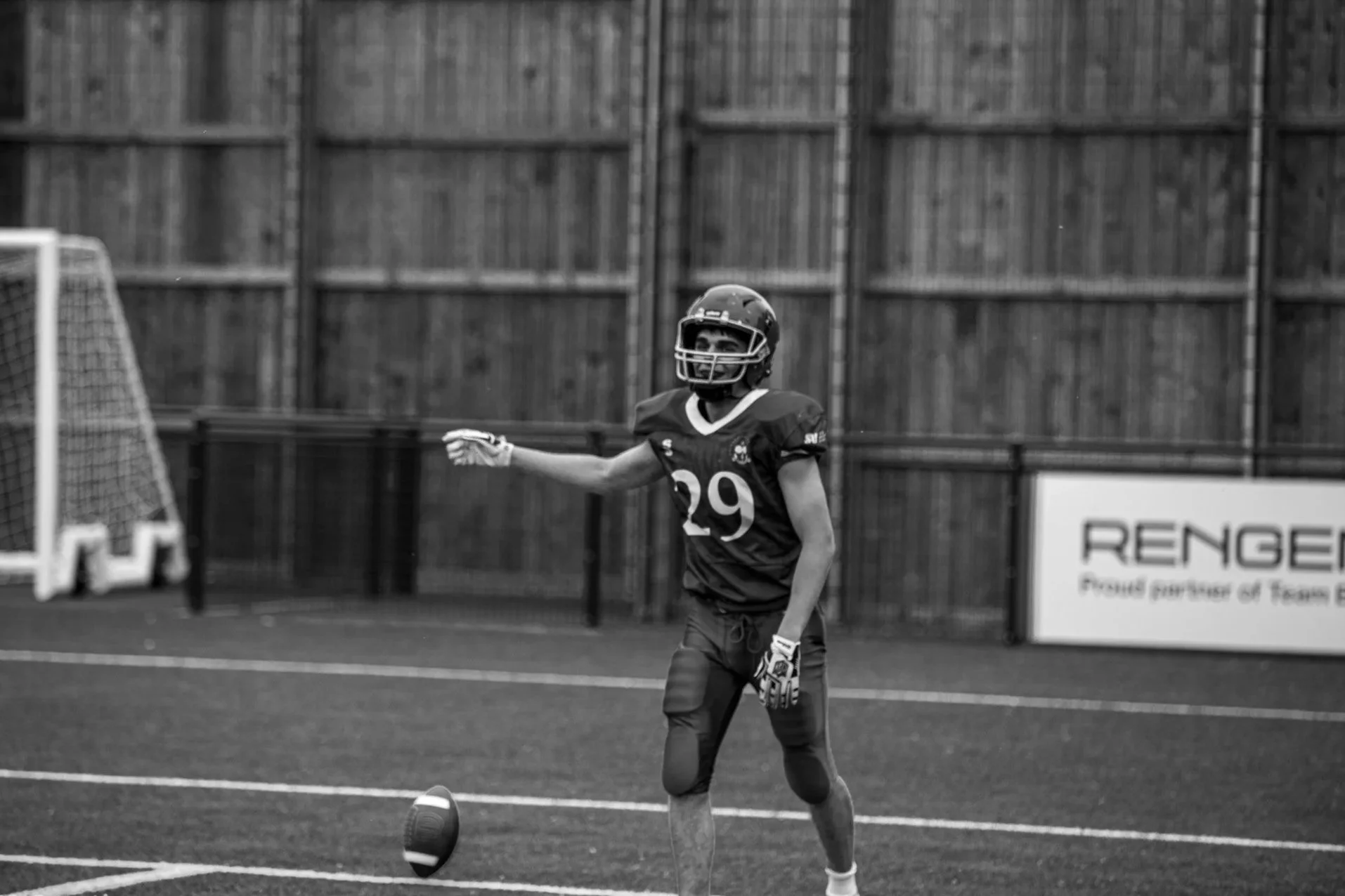 A young football player wearing a helmet, jersey, and gloves is on a field, mid-action, with a football at his feet and an encouraging expression, near a goalpost and a fence in the background.