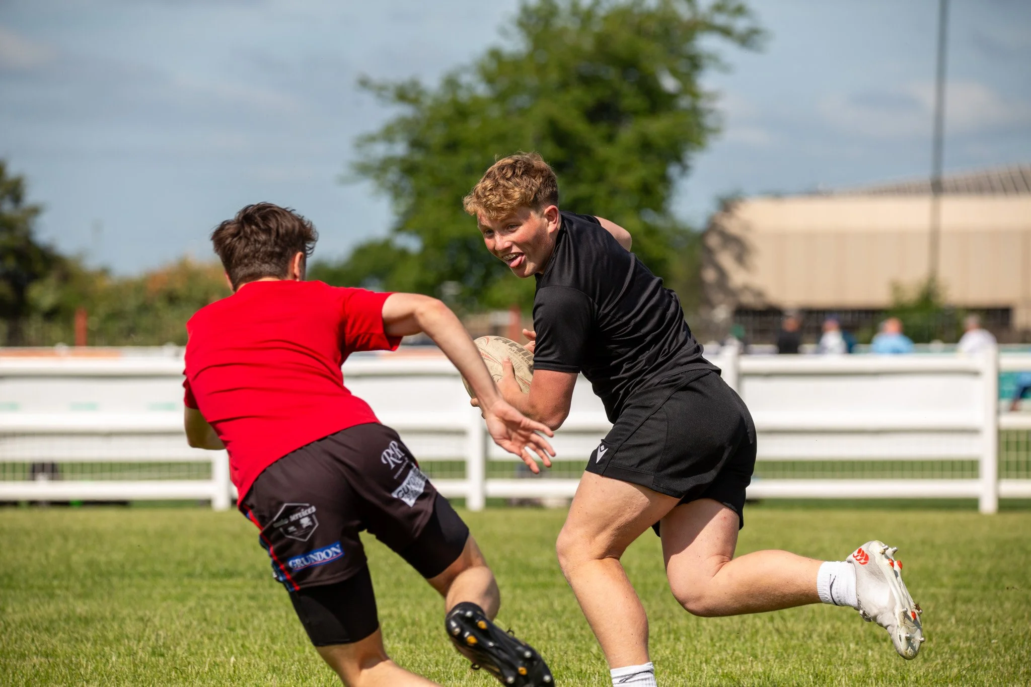 Two boys playing rugby outside on a sunny day, with one boy holding a rugby ball and both wearing sportswear, running on a grassy field.