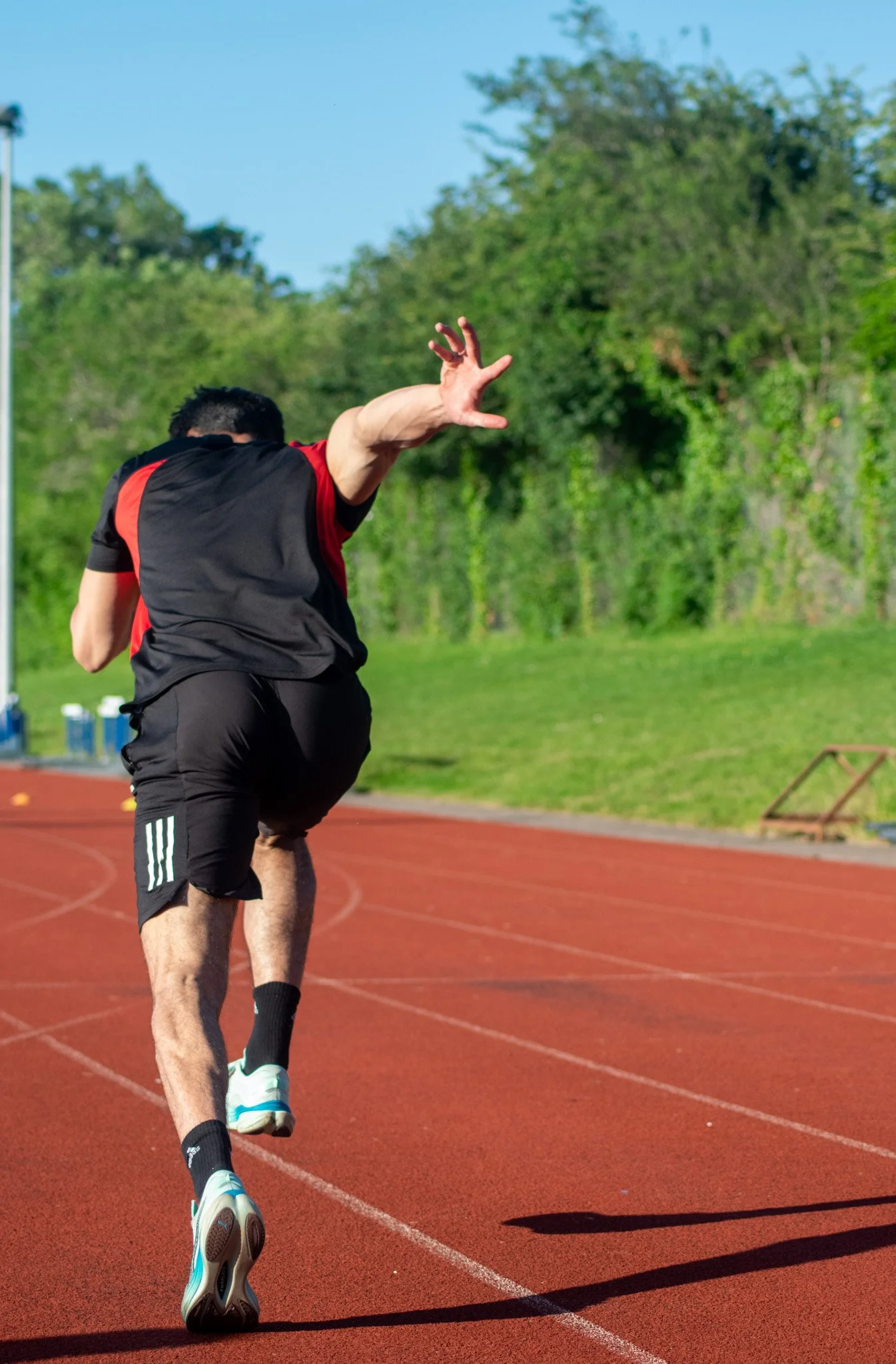A person in sports attire running on an outdoor track during daytime, with trees and a clear blue sky in the background.