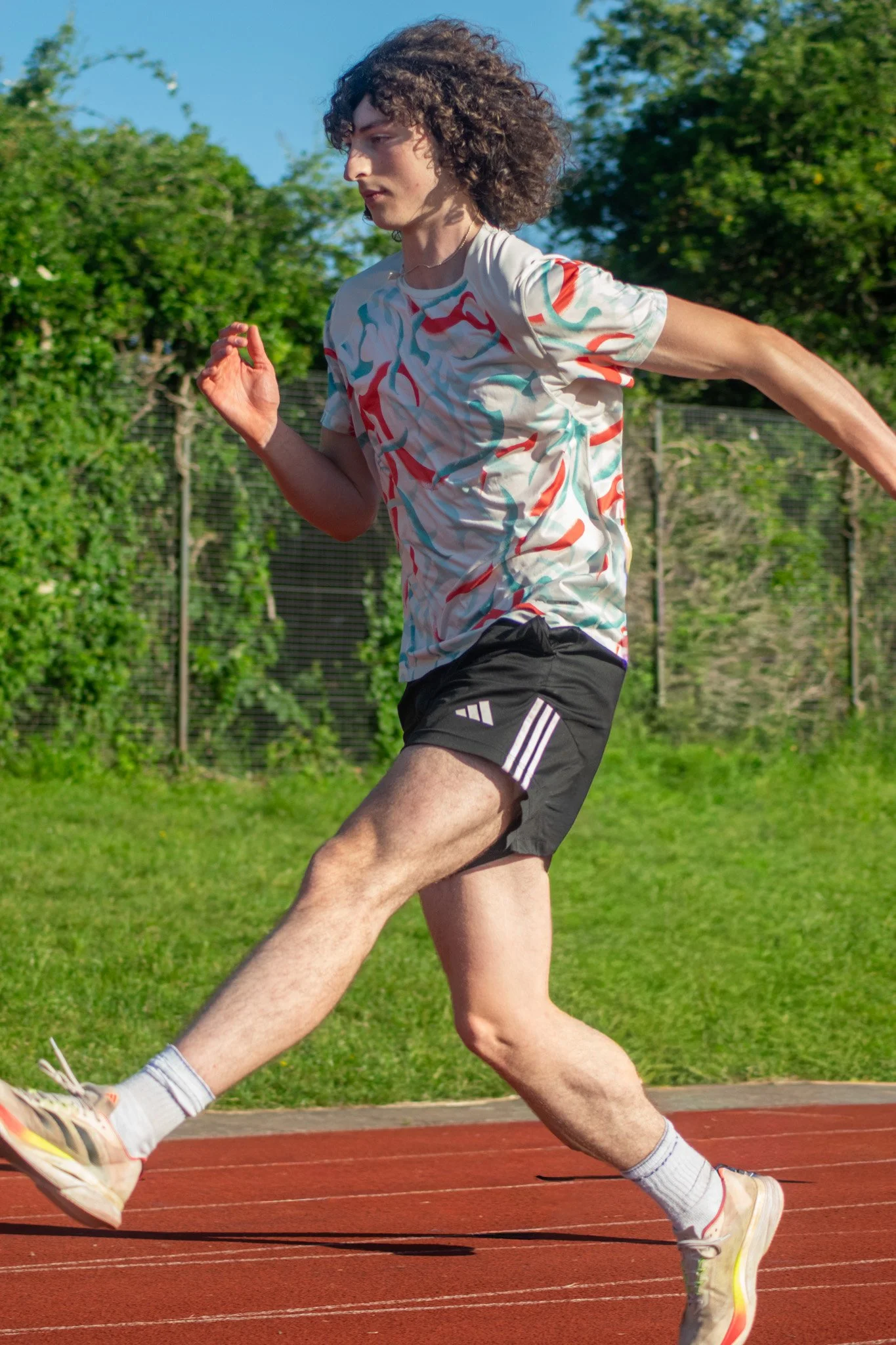 A man running on a red outdoor track field, wearing a white shirt with colorful abstract patterns, black shorts, white socks, and running shoes, with a background of green trees and a clear blue sky.