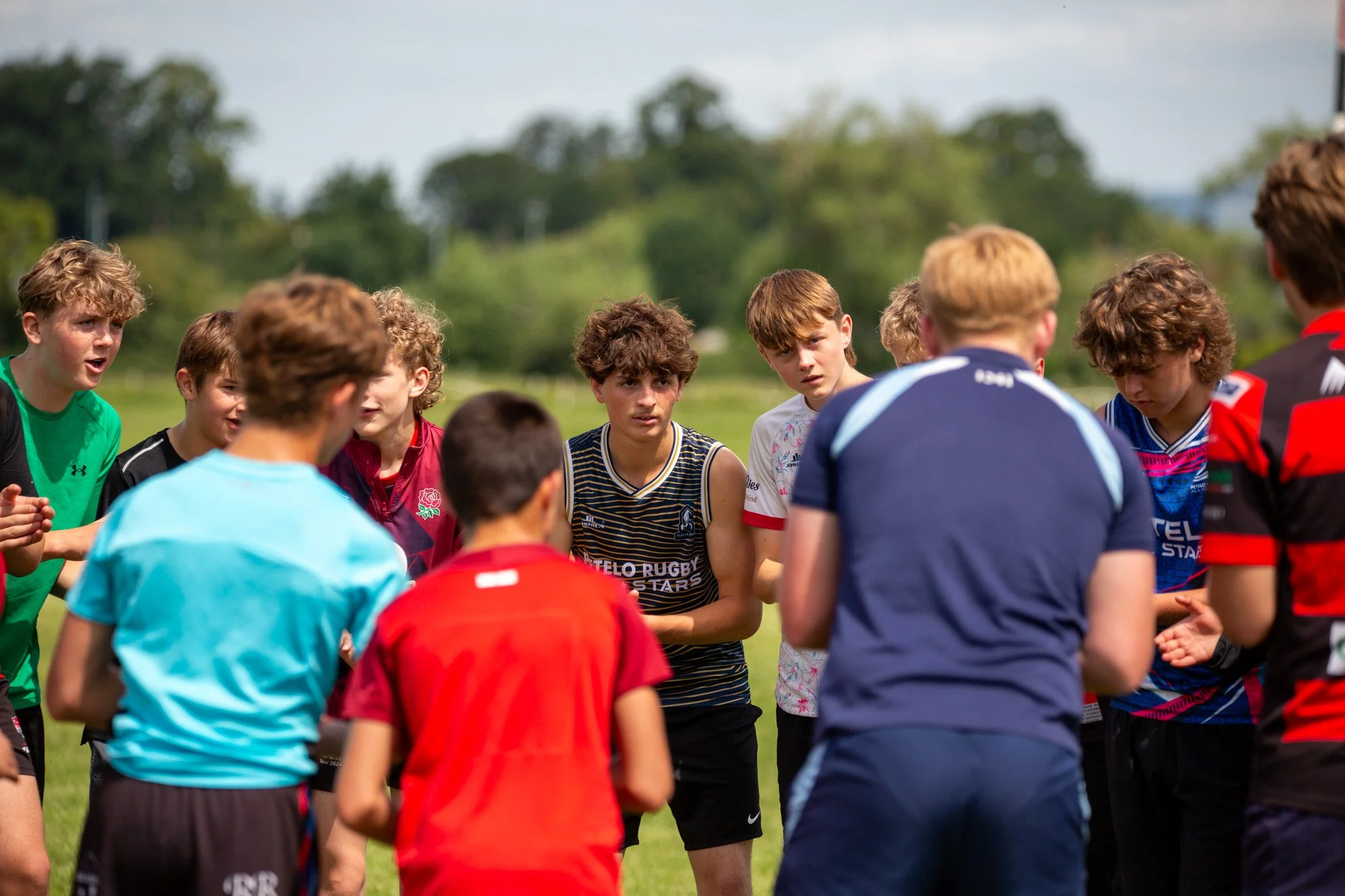 A group of young boys gathered outdoors on a grassy field during daytime, dressed in sports attire, likely participating in a rugby practice or game, with trees and a cloudy sky in the background.