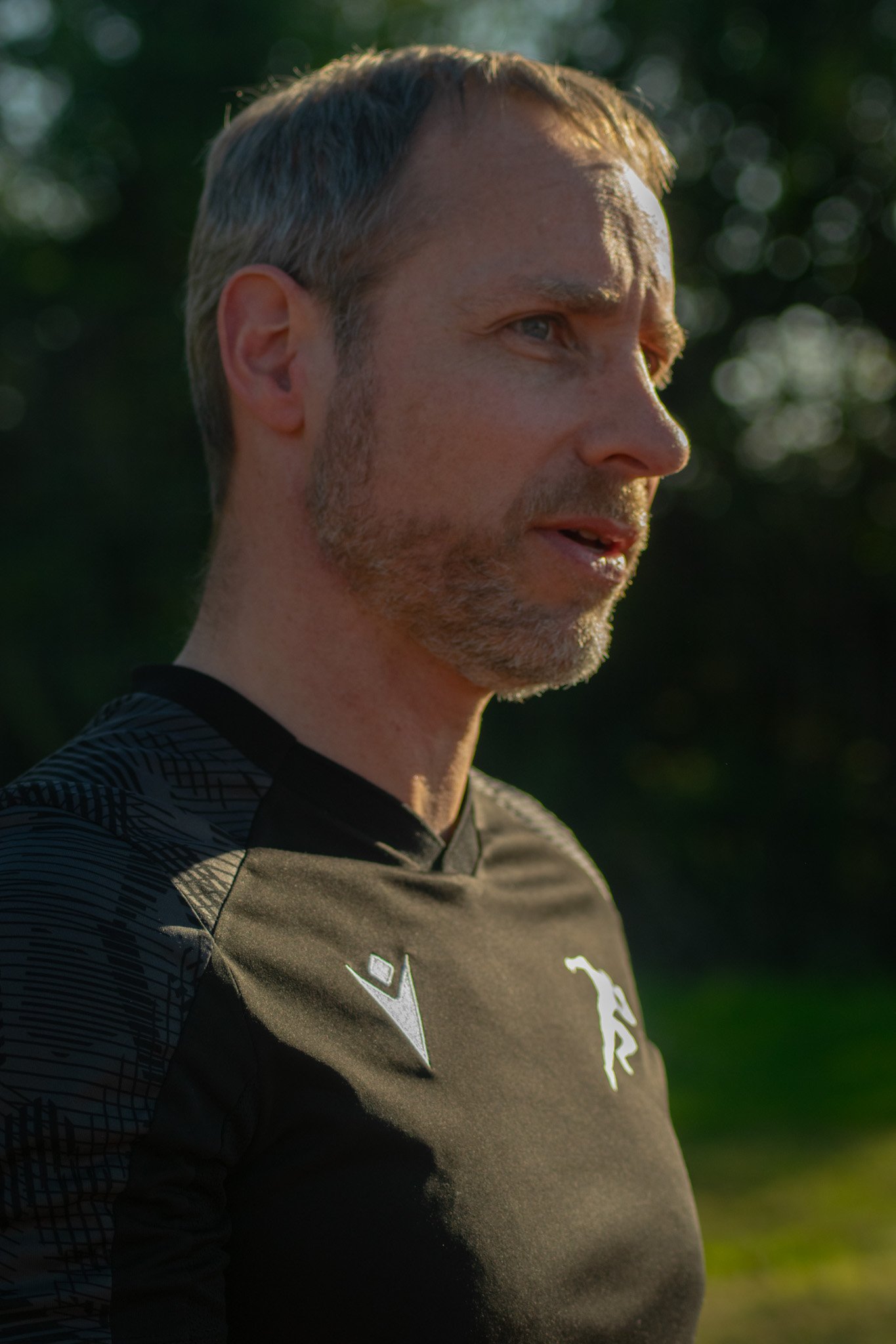 Close-up of a middle-aged man with short gray hair and light stubble, wearing a black athletic shirt, outdoors with blurred trees in the background.
