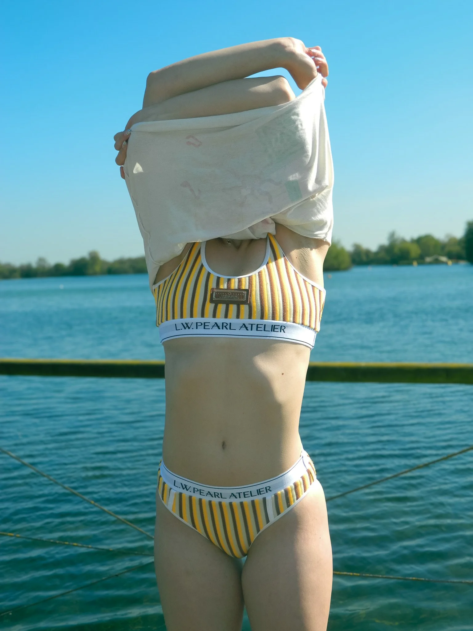 A person standing outdoors near a body of water, holding a white shirt over their head, with a clear blue sky above and some trees visible in the background.