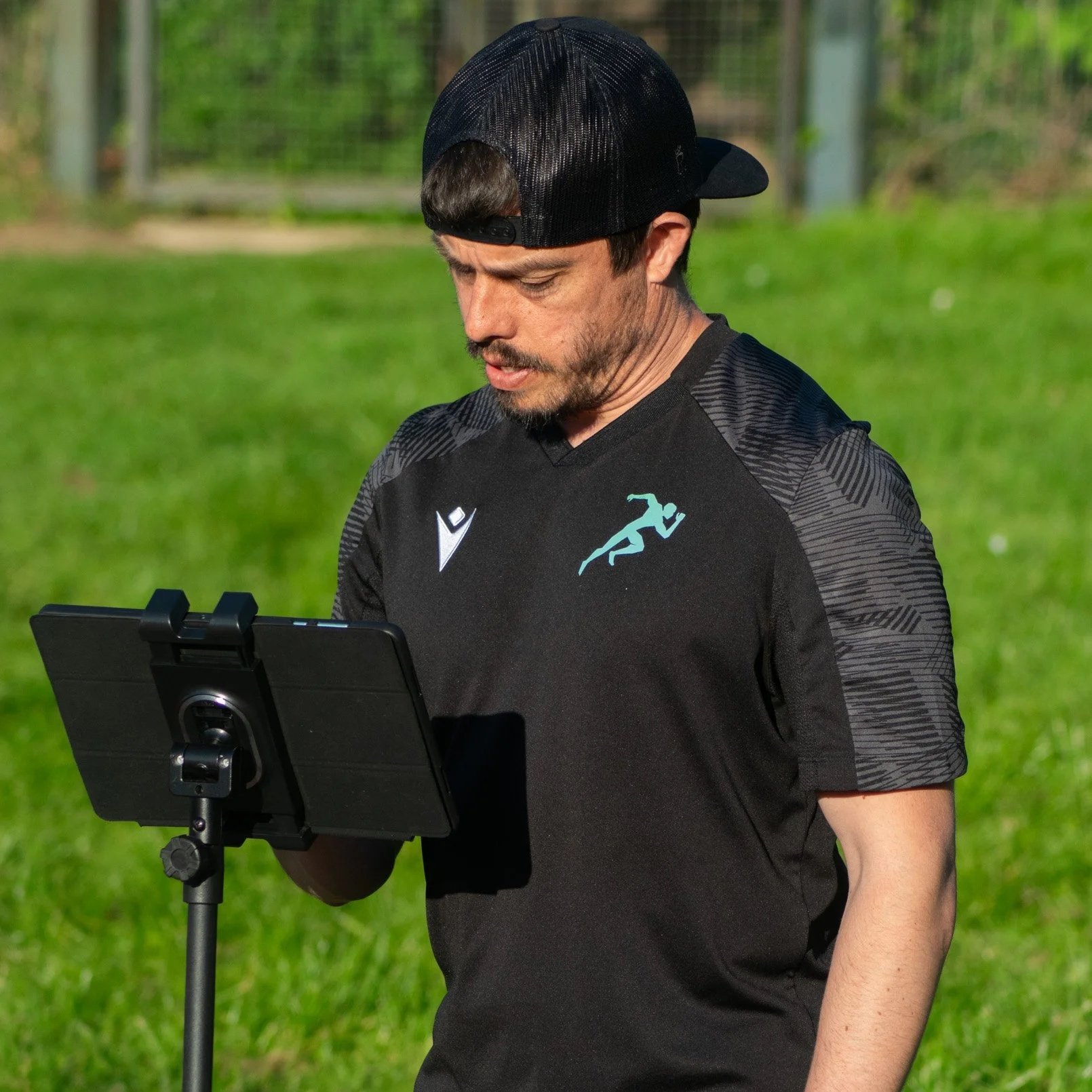A man wearing a black baseball cap and a black sports shirt with a logo, standing outdoors on grass, looking at a tablet mounted on a stand.