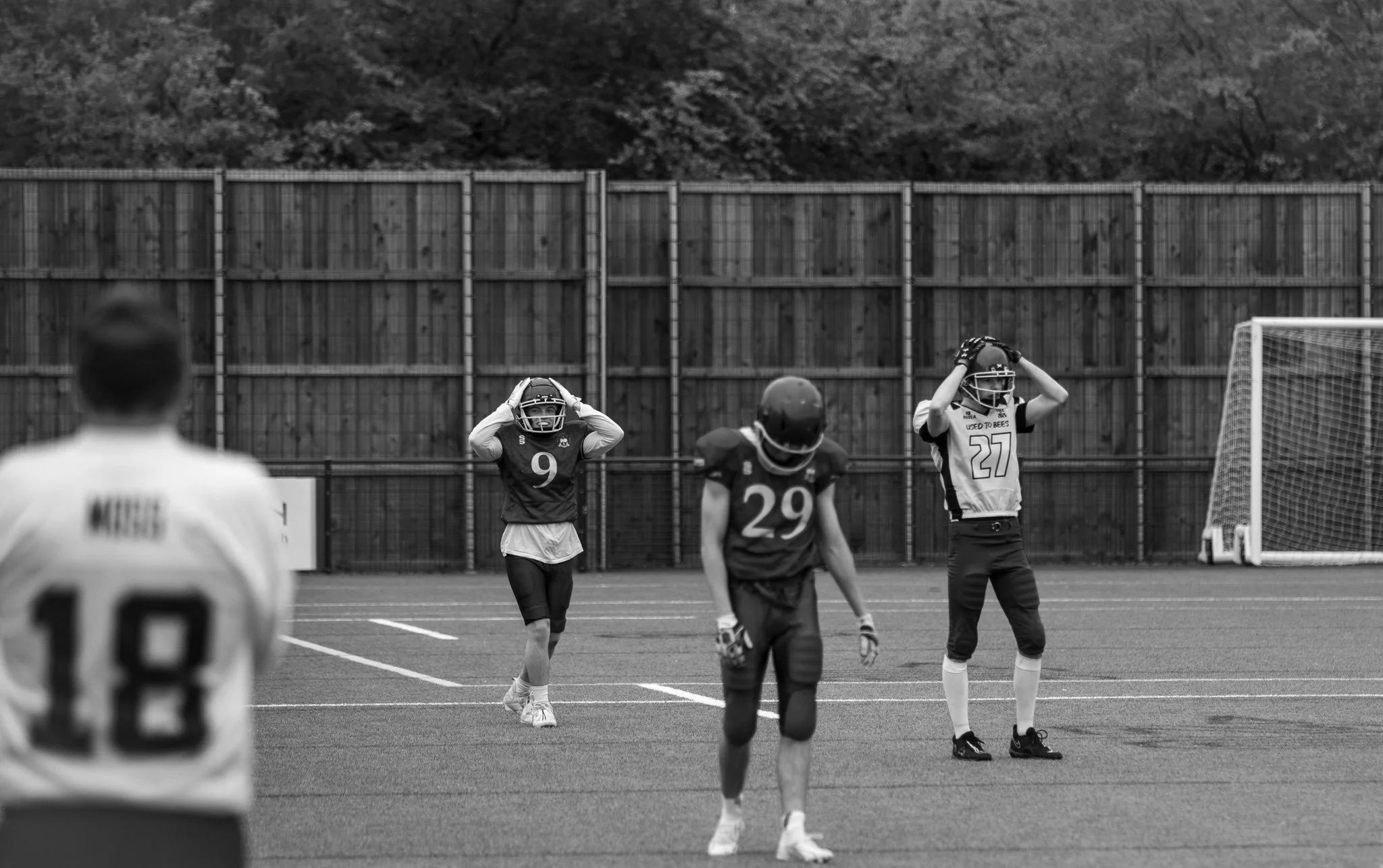 A group of football players on the field, some with hands on their helmets, standing near the goalpost during a game in daytime, with a wooden fence and trees in the background.