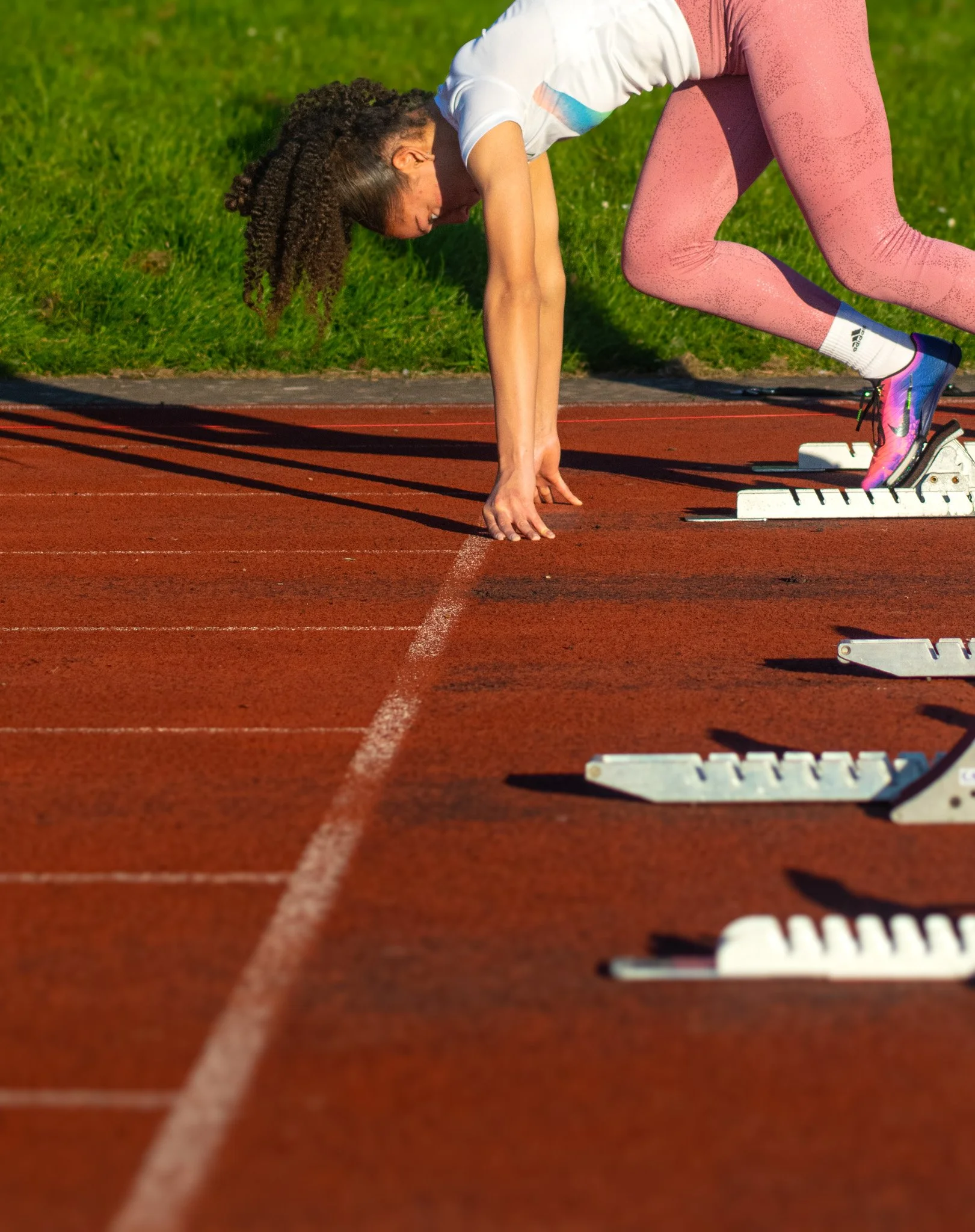 A woman in pink leggings and a white t-shirt preparing for a sprint on a starting block on a red running track.